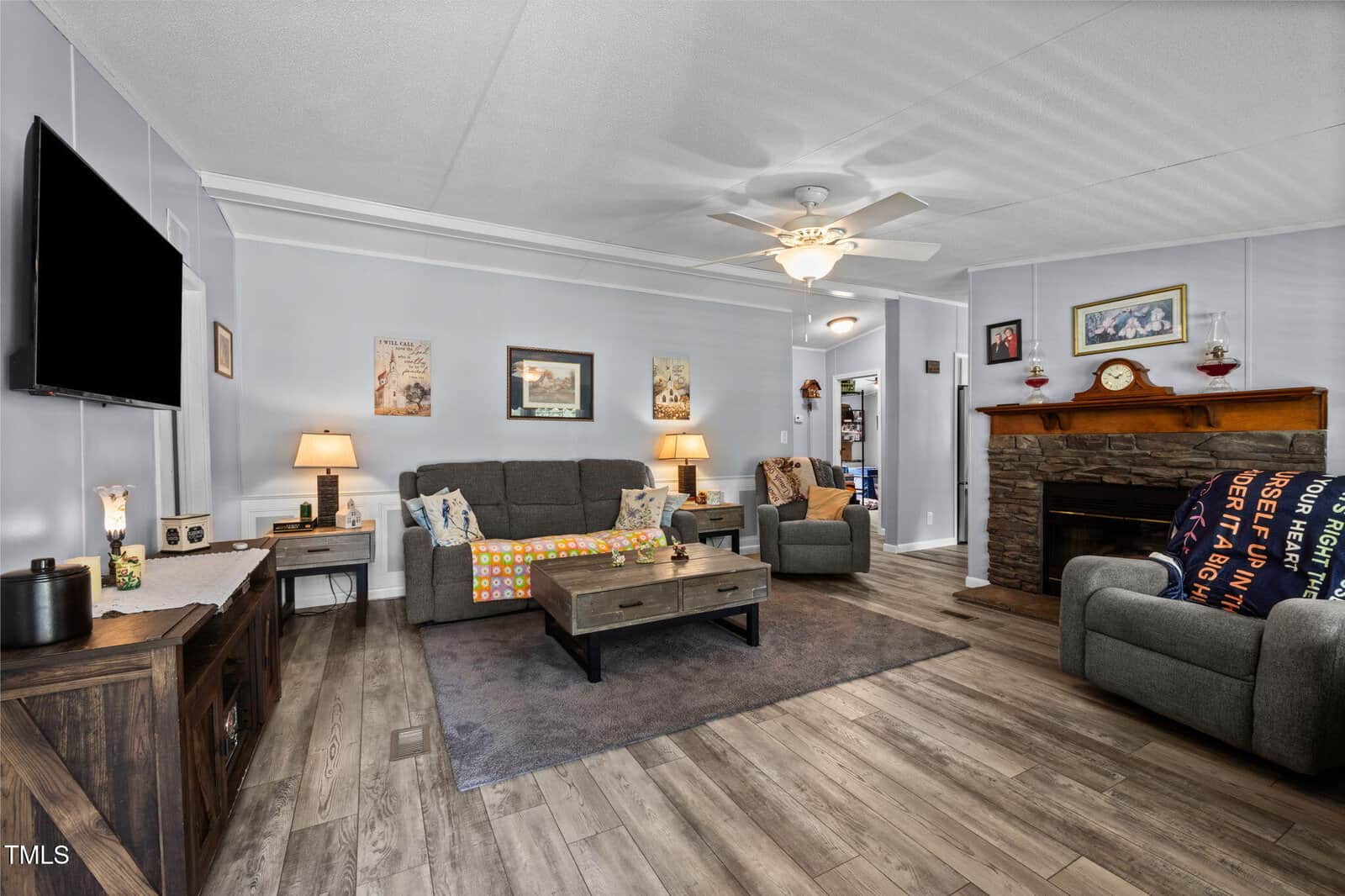 A cozy living room with gray walls, wood flooring, a gray sofa, armchair, TV, coffee table, fireplace with mantle, lamps, and colorful blankets. Art and photos hang on the walls.