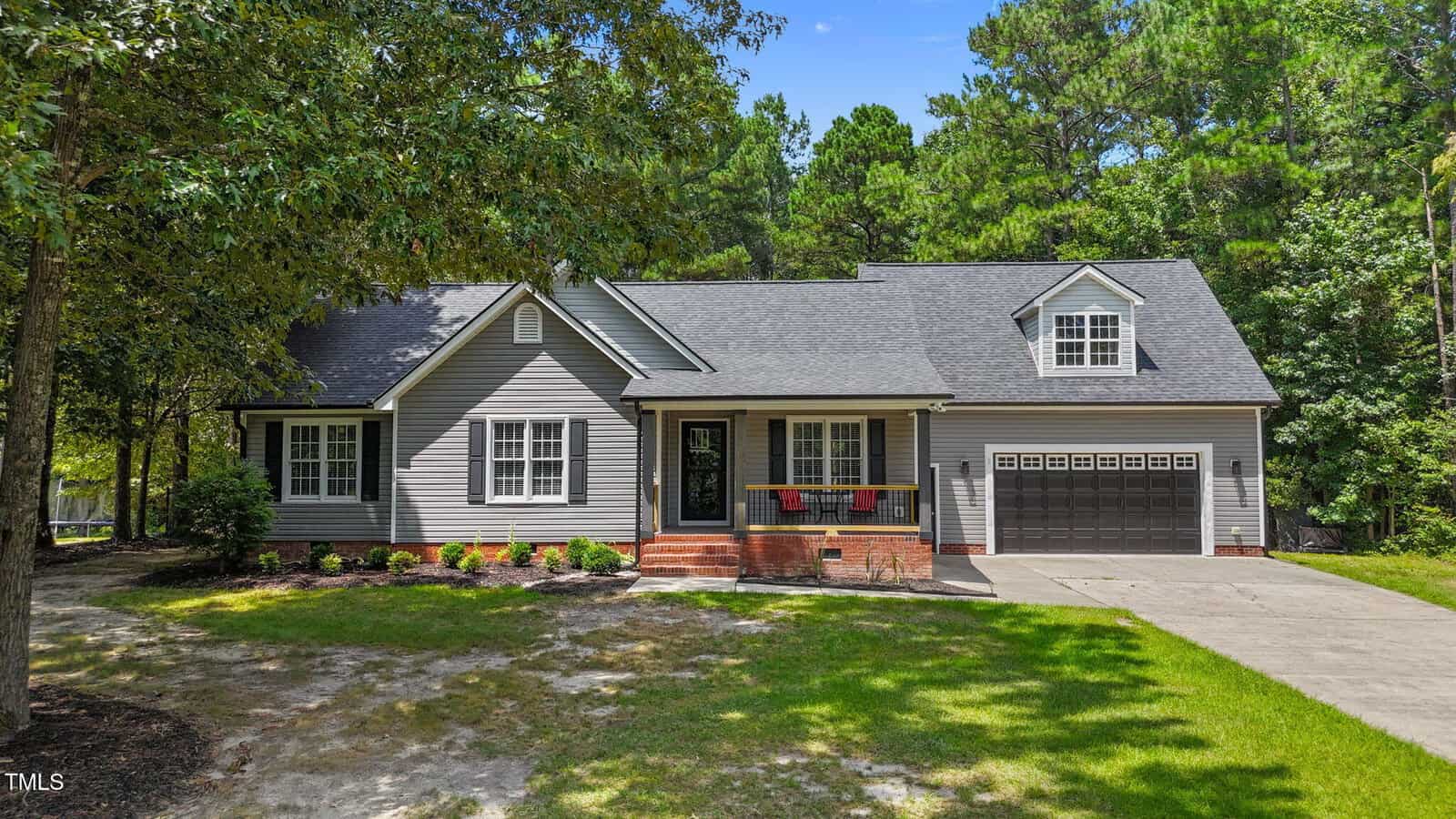 A gray suburban house with a gabled roof, black shutters, and a two-car garage sits surrounded by trees and green lawn under a clear blue sky. Brick steps lead up to the front door and flower boxes hang under the windows.