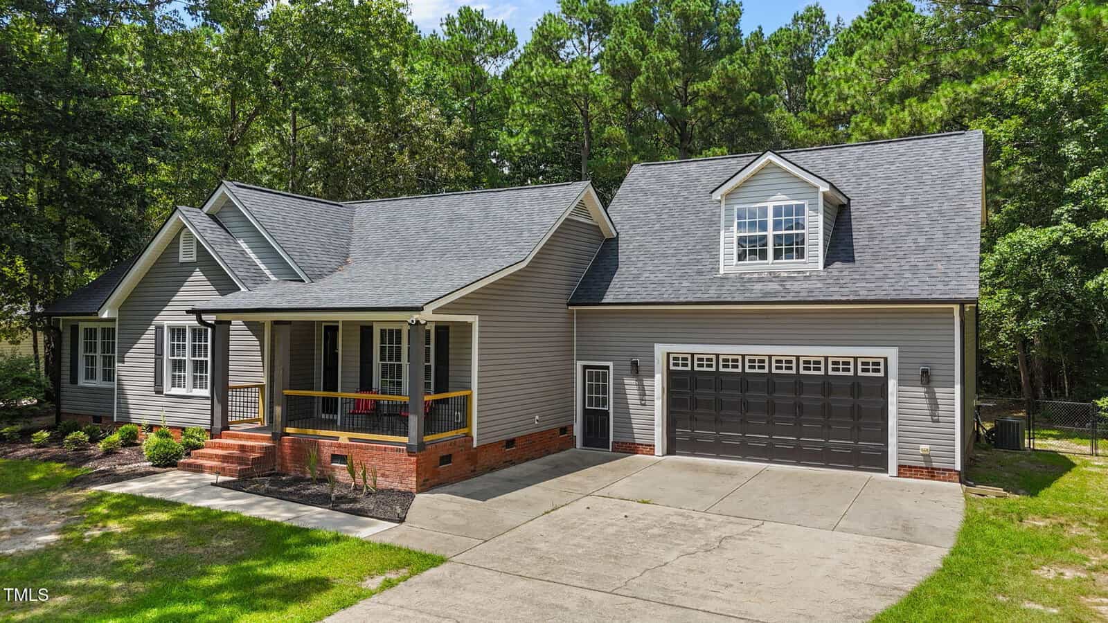 A modern gray house with white trim, a covered front porch, and a double garage. The home sits on a concrete driveway and is surrounded by green trees and landscaped yard.