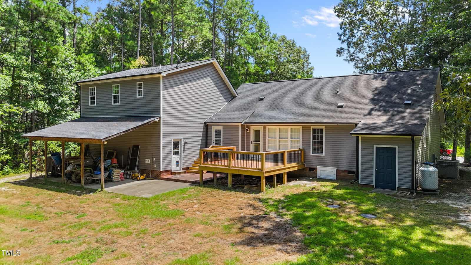 A two-story house with gray siding, a covered carport, wooden deck, back entrance, and a yard bordered by trees under a blue sky.