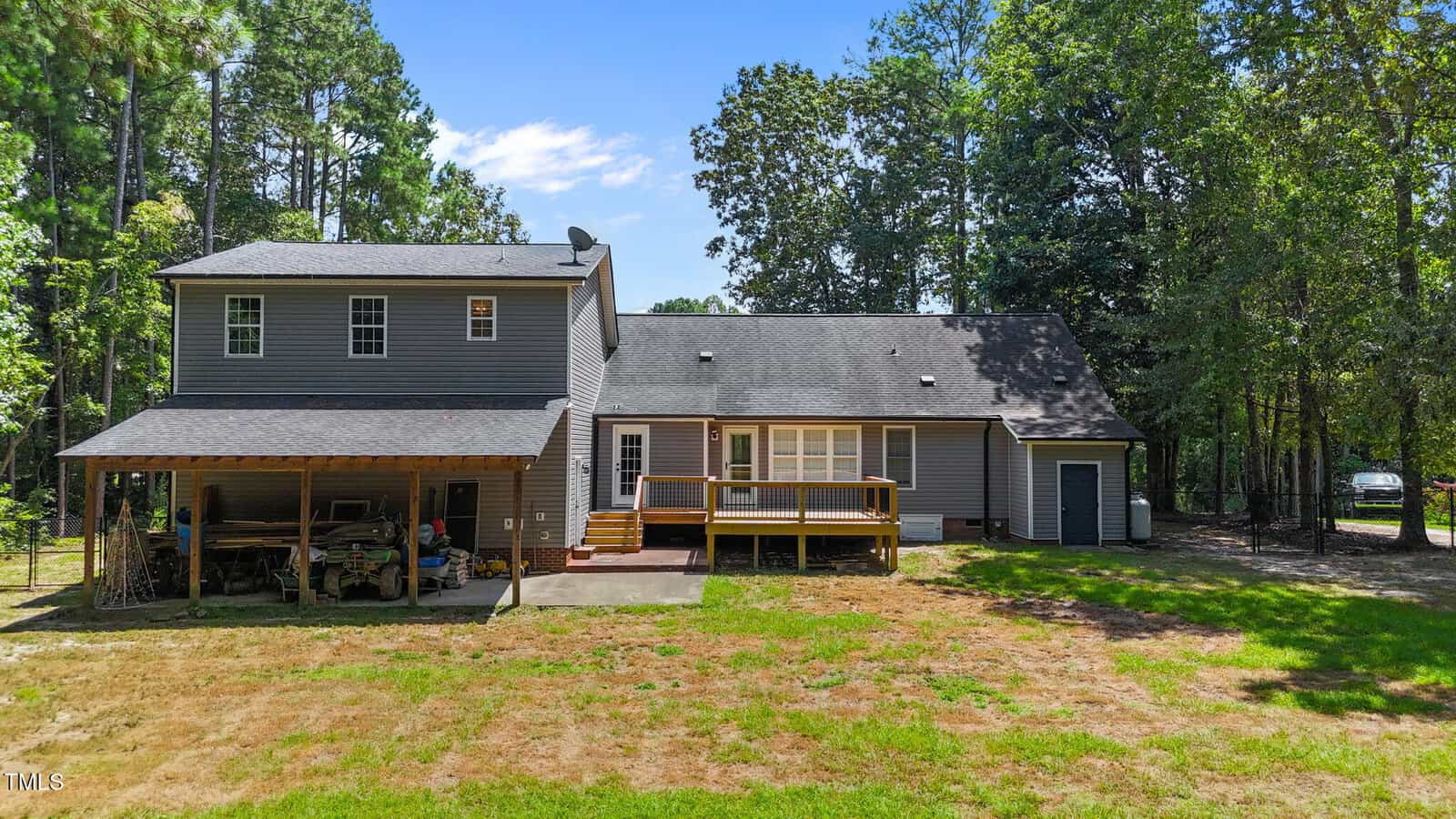 A two-story gray house with a covered carport on the left and a wooden deck at the back, surrounded by trees and a grassy yard under a clear blue sky.