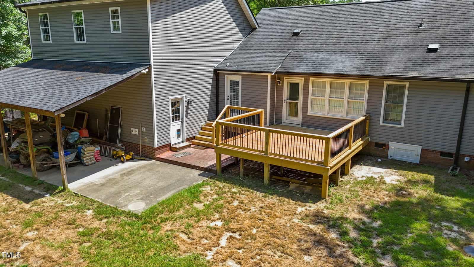 A gray house with a wooden deck attached to the back, beside a covered carport with stored tools and items. The yard is patchy with grass and dirt. Steps lead from the deck to the ground.