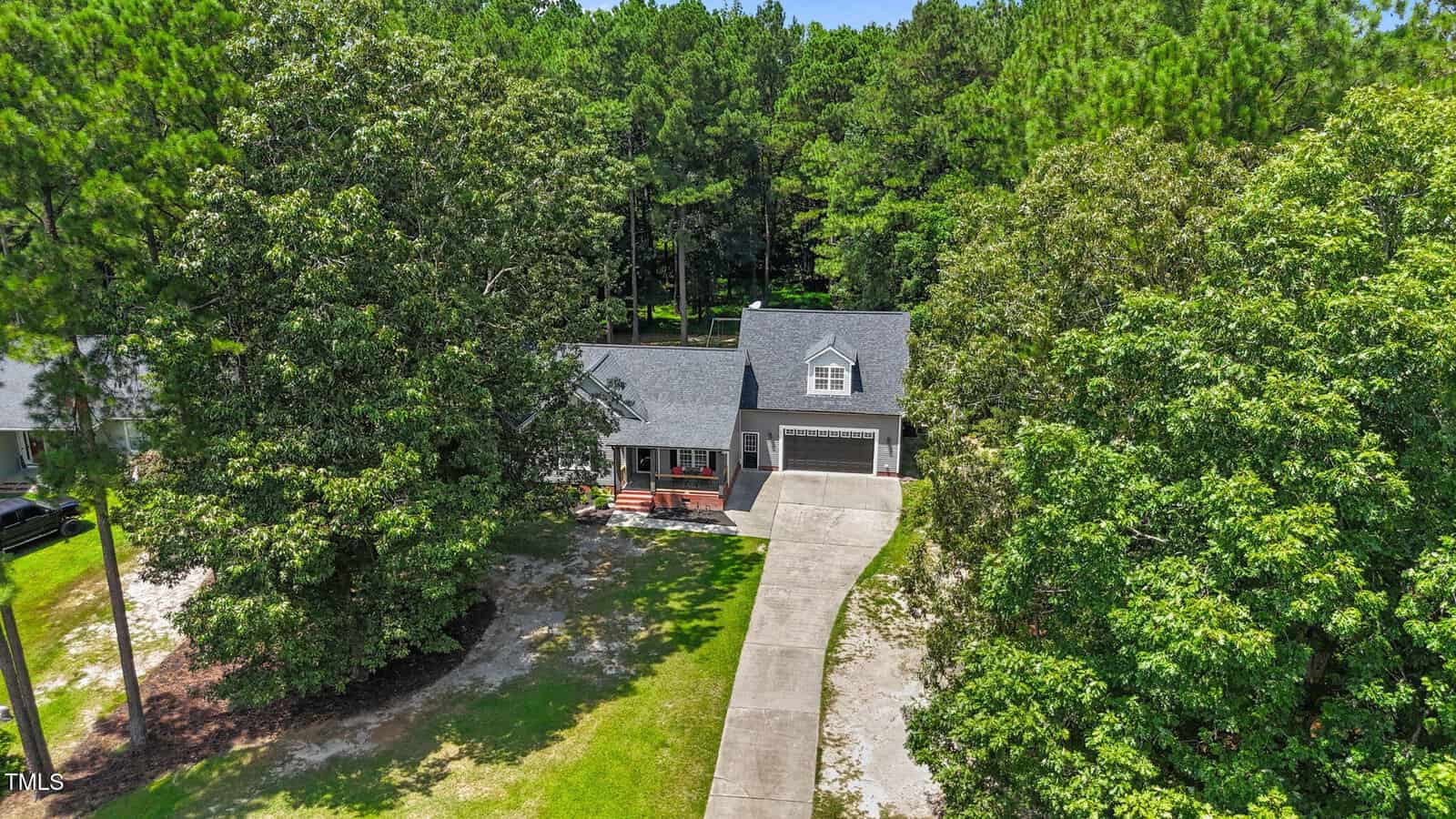 A gray house with a front porch and attached garage sits among tall green trees. A curved concrete driveway leads to the garage. The yard is partly shaded and bordered by a forested area.
