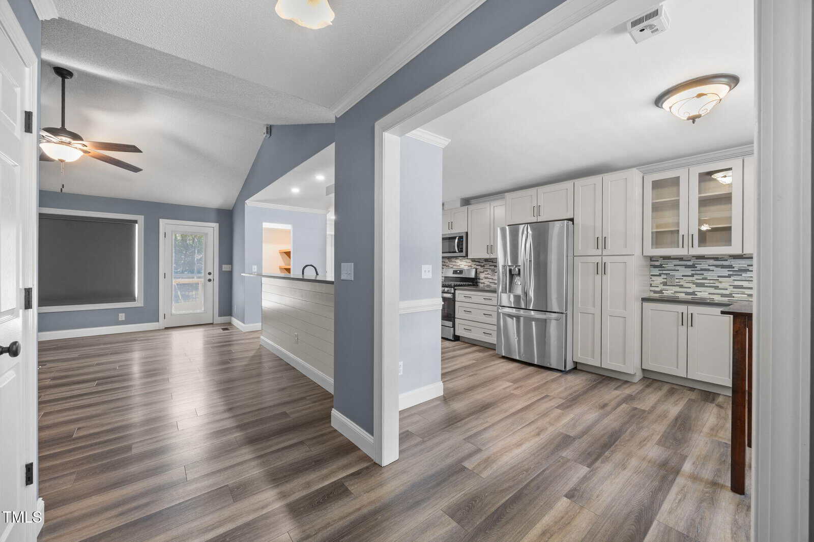 Open kitchen and dining area with stainless steel appliances, white cabinets, light wood flooring, gray and blue walls, ceiling fans, and large windows. Modern design with a bright, clean look.