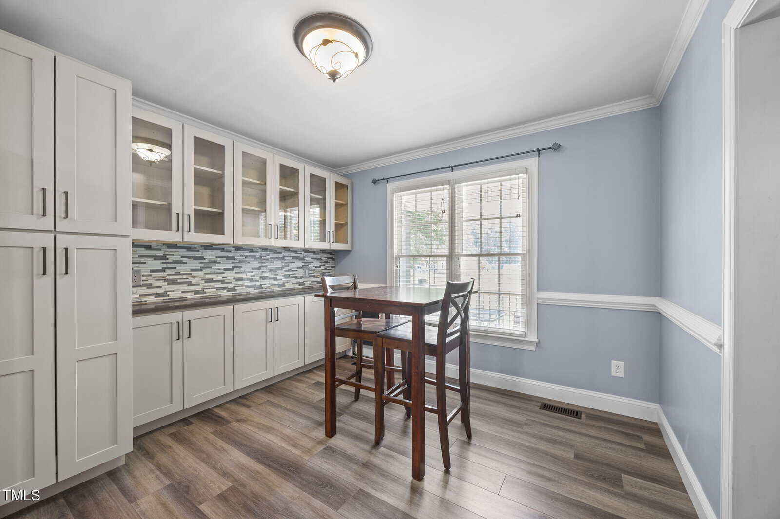 A kitchen area with light blue walls, white cabinets with glass doors, a tile backsplash, and a small wooden dining table with two chairs in front of a window with blinds. The floor is wood laminate.