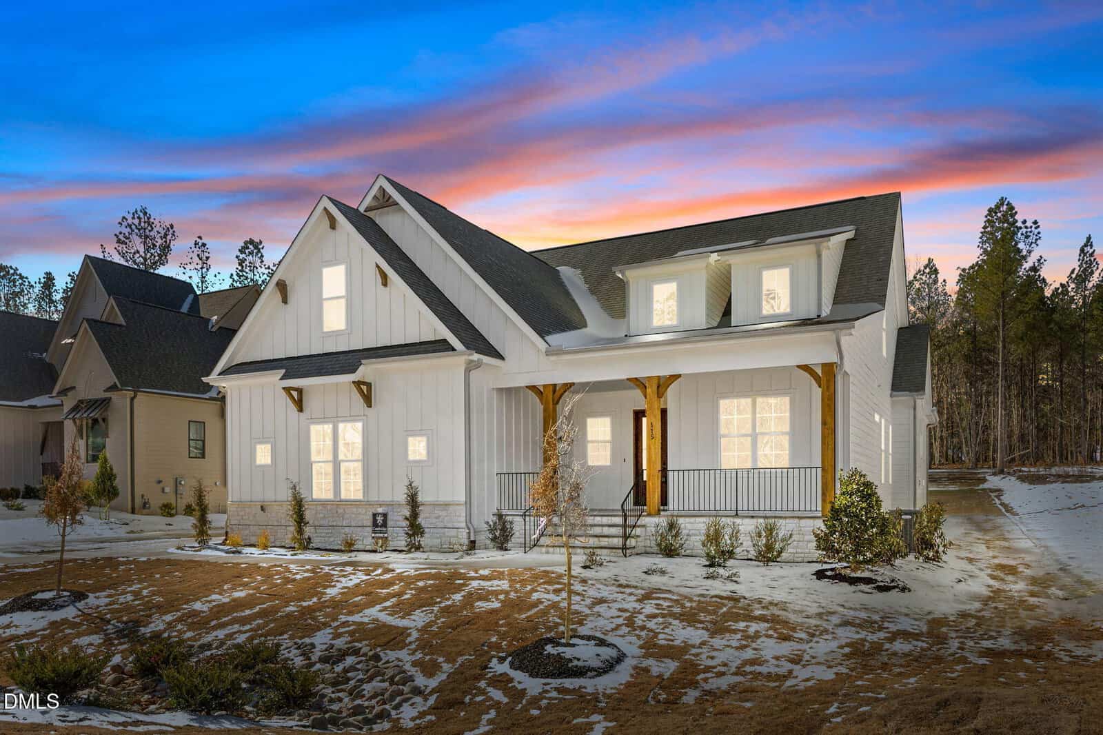 A modern white house with a dark roof and front porch, illuminated warmly at dusk at 115 Cherry Bark Drive, Youngsville. Snow blankets the ground and roof beneath a colorful sunset sky, framed by trees in the background.