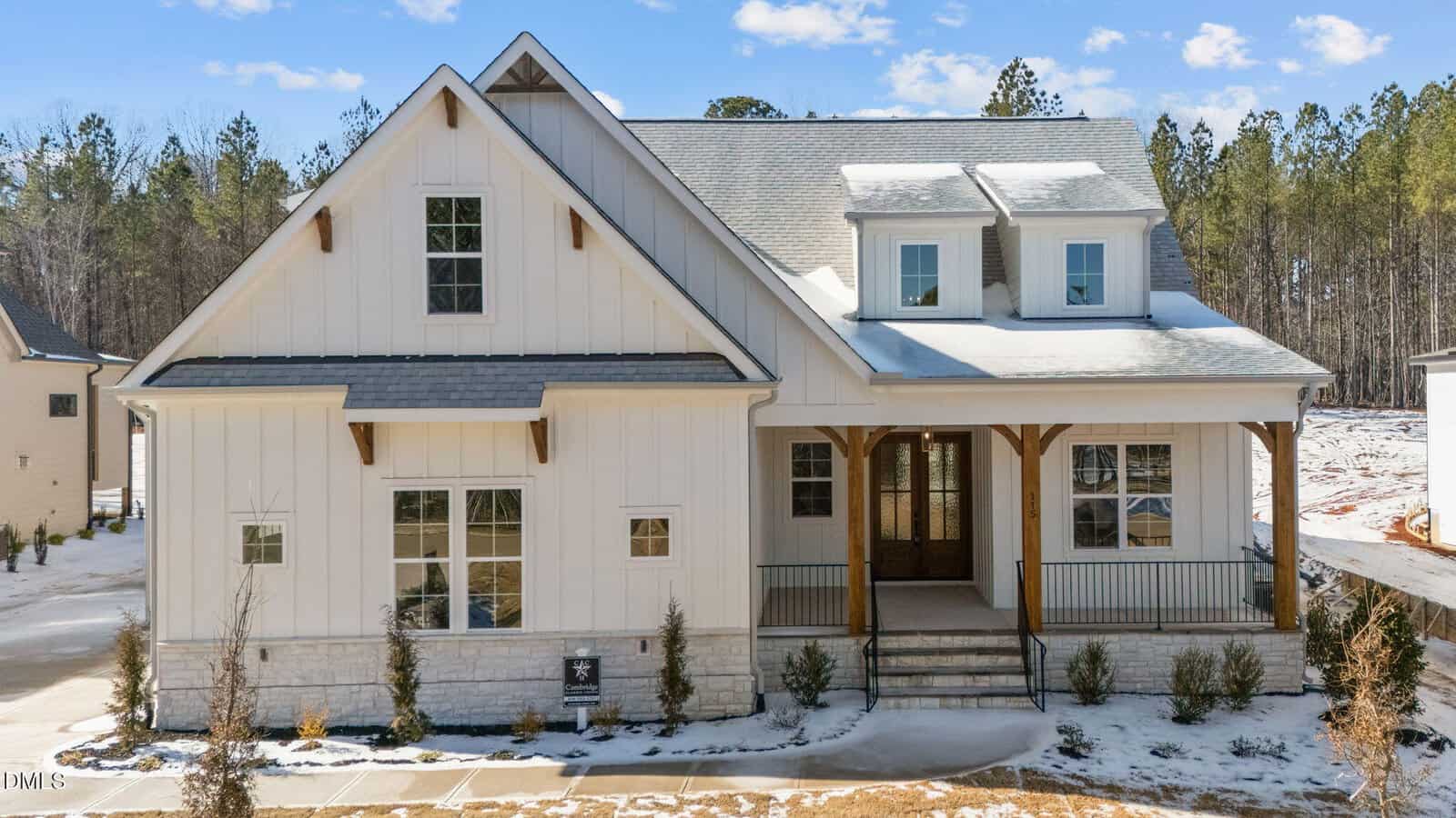 A modern farmhouse with white siding, dark roof shingles, front porch with wooden beams, and dormer windows. The house is surrounded by light snow and has a wooded area in the background.