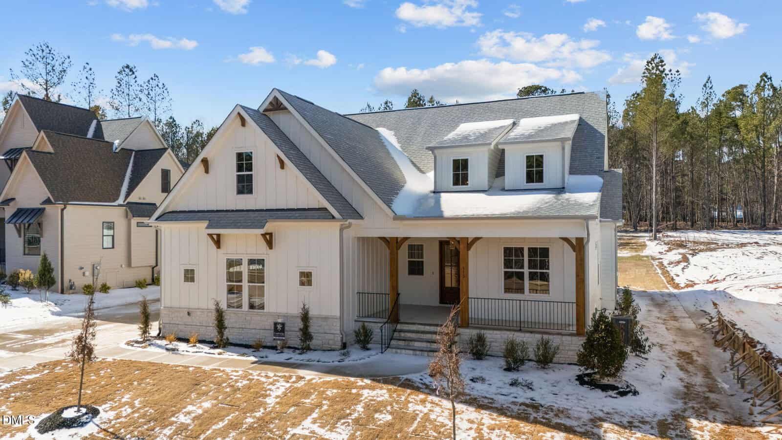 A modern white house with a covered front porch, gabled roof, and dormer windows sits on a snowy yard with young trees, surrounded by neighboring homes and a background of tall pine trees.