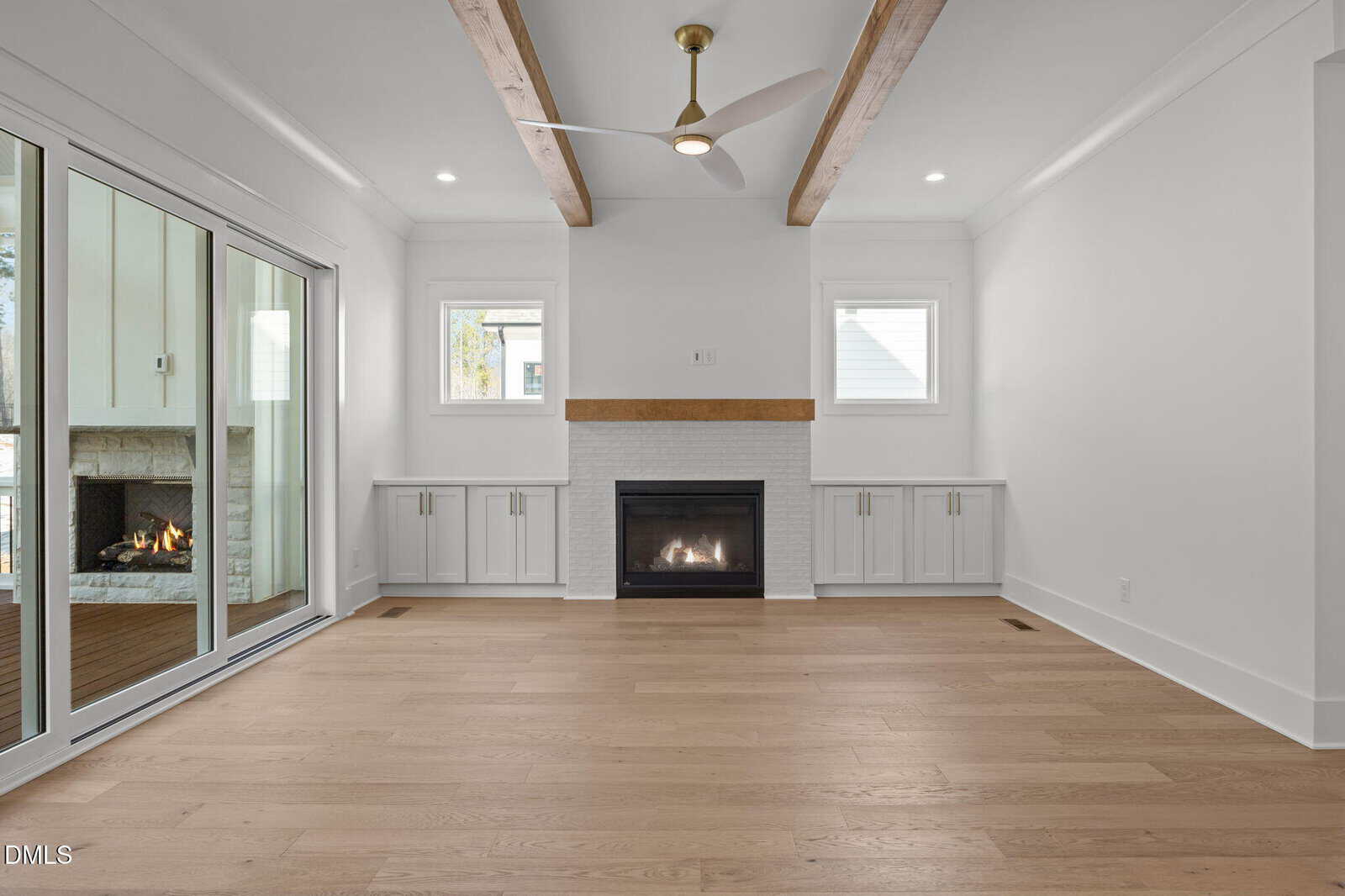 Modern living room with light wood floors, a white brick fireplace, built-in cabinets, wood ceiling beams, and a ceiling fan. Large sliding glass doors open to an outdoor patio with a fireplace.