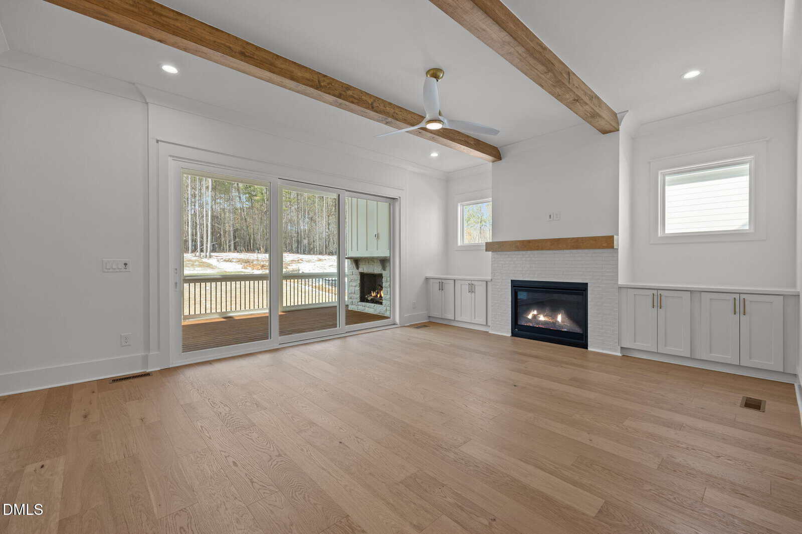Modern living room with light wood floors, white walls, exposed wooden ceiling beams, built-in cabinets, a gas fireplace, ceiling fan, and large sliding glass doors opening to a deck with a view of trees.