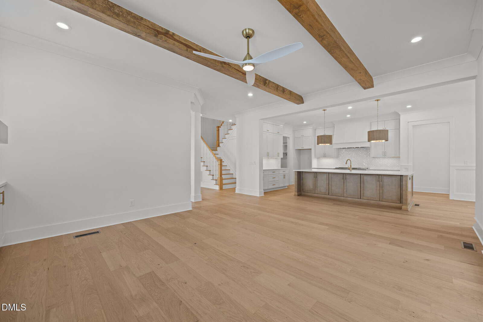 Spacious open-concept kitchen and living area with wood floors, white walls, exposed wood ceiling beams, a ceiling fan, modern lighting, and a staircase in the background.