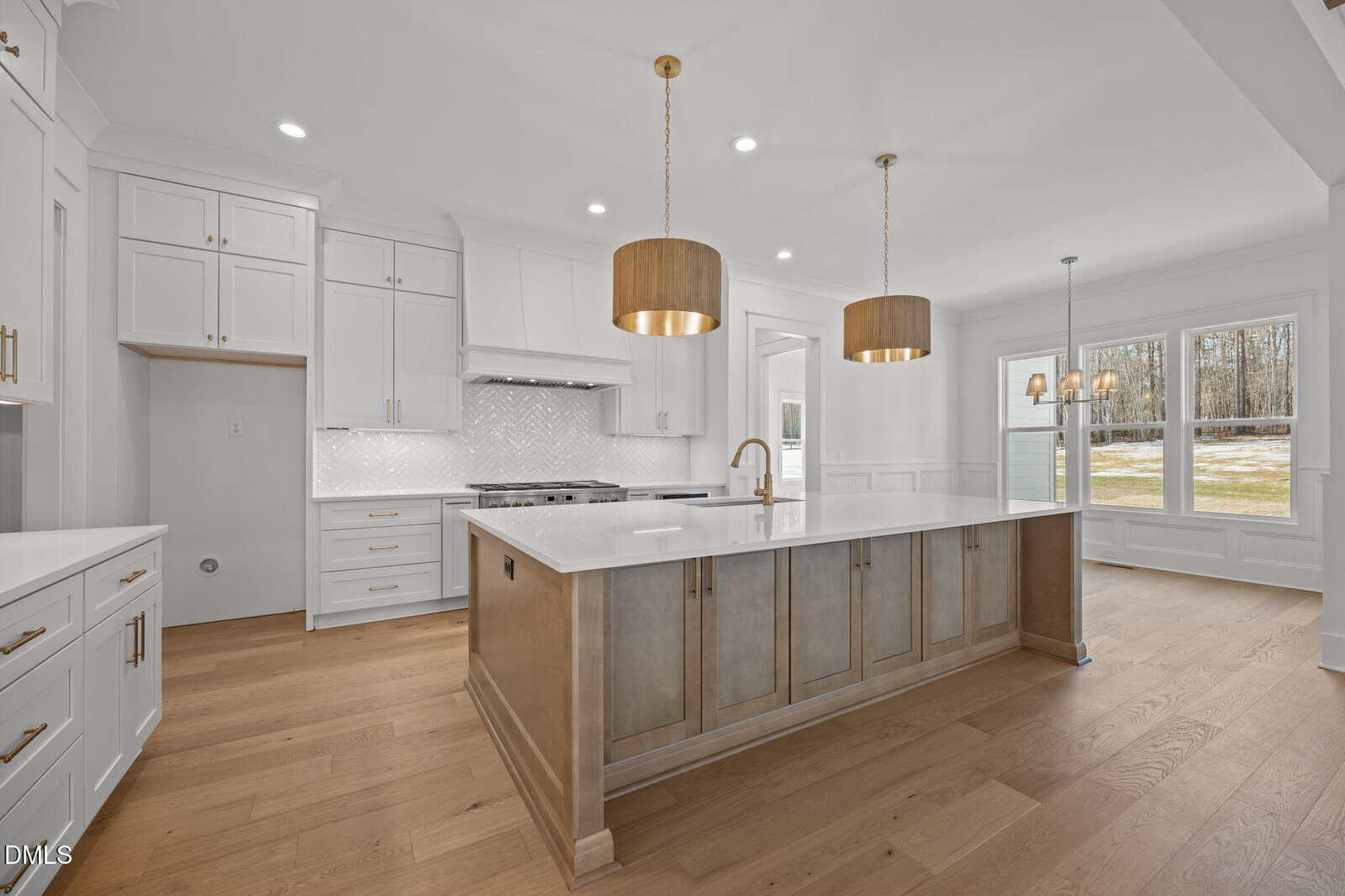 Modern kitchen with white cabinets, a large center island with a wood finish, gold pendant lights, stainless steel appliances, and wide plank hardwood floors. Large windows let in natural light, offering views of the outdoors.