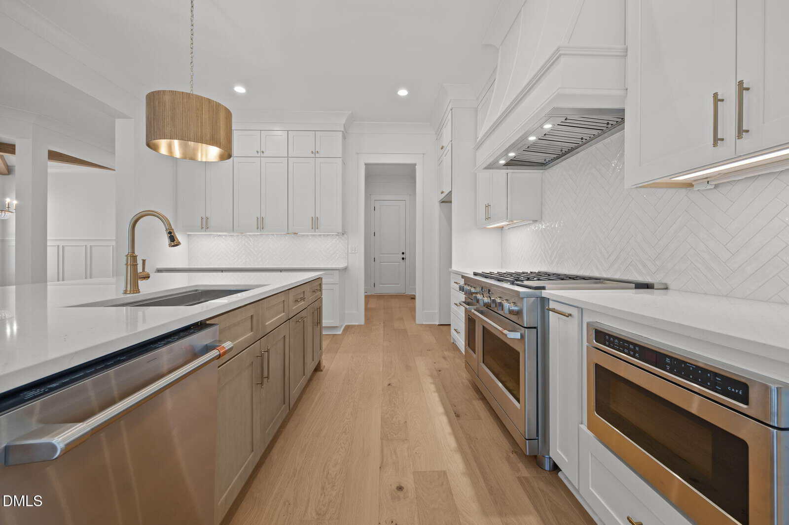 Modern kitchen with white cabinets, stainless steel appliances, gold fixtures, an island with sink, wood floor, and herringbone tile backsplash. A gold pendant light hangs above the island.