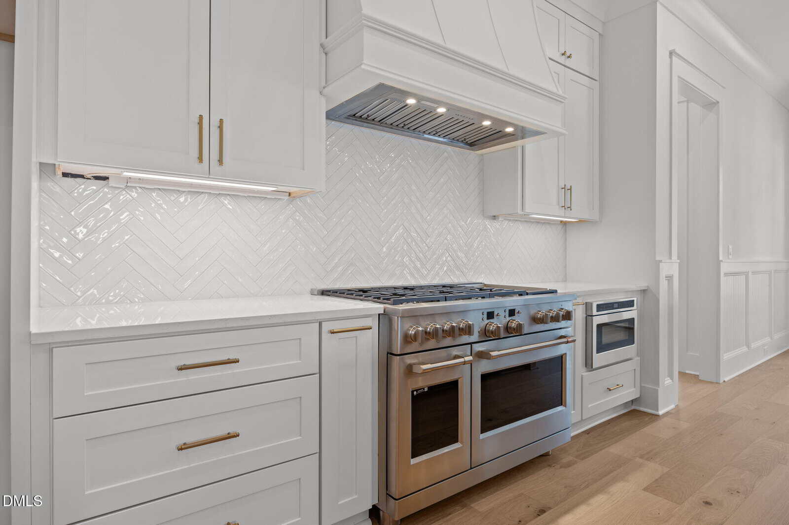 Modern kitchen with white cabinets, gold handles, stainless steel double oven and stove, under-cabinet lighting, light wood floor, and a white herringbone tile backsplash.
