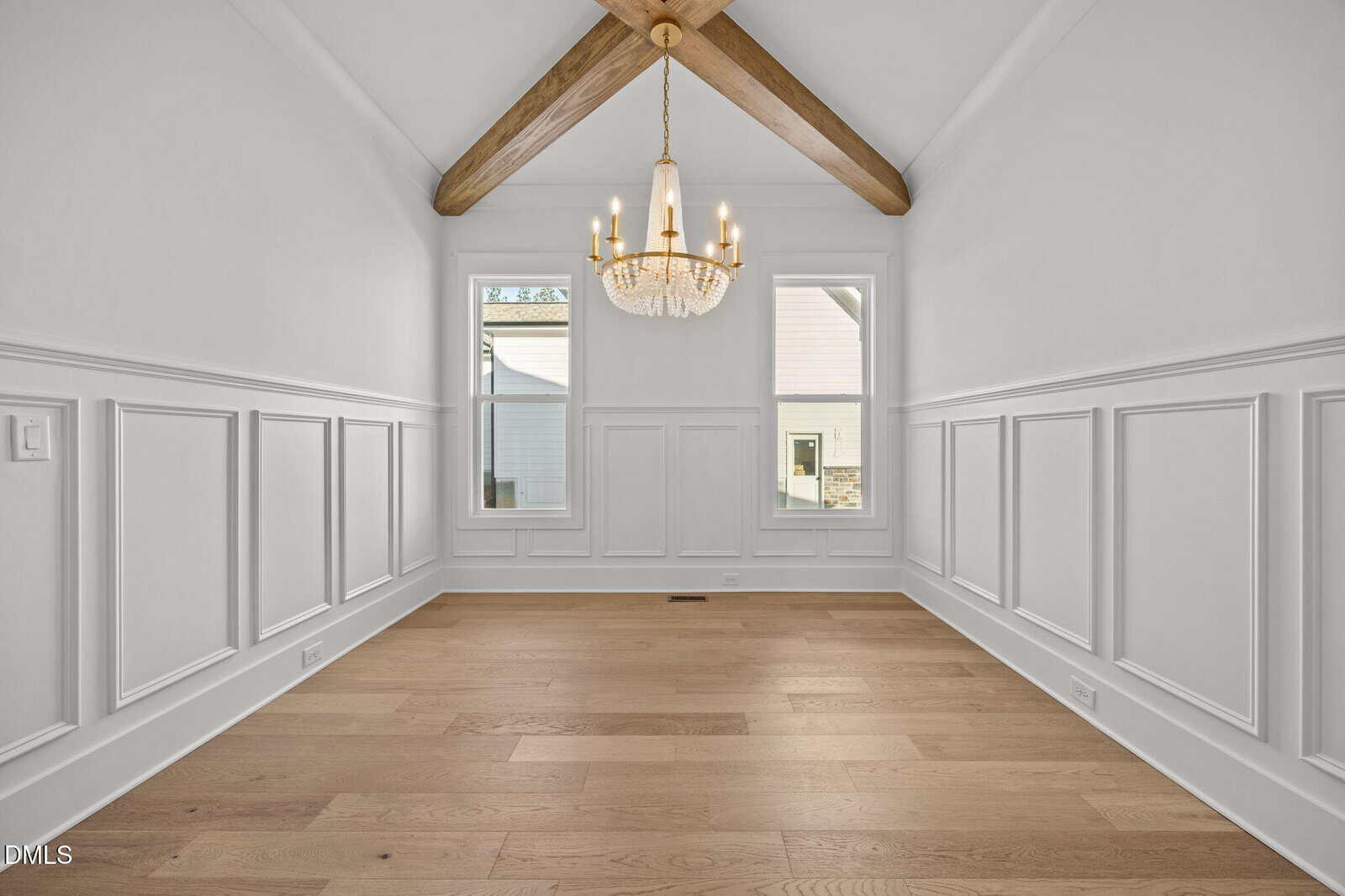 A bright, empty room with white paneled walls, light wood floors, two tall windows, exposed wood ceiling beams, and a chandelier hanging from the vaulted ceiling.