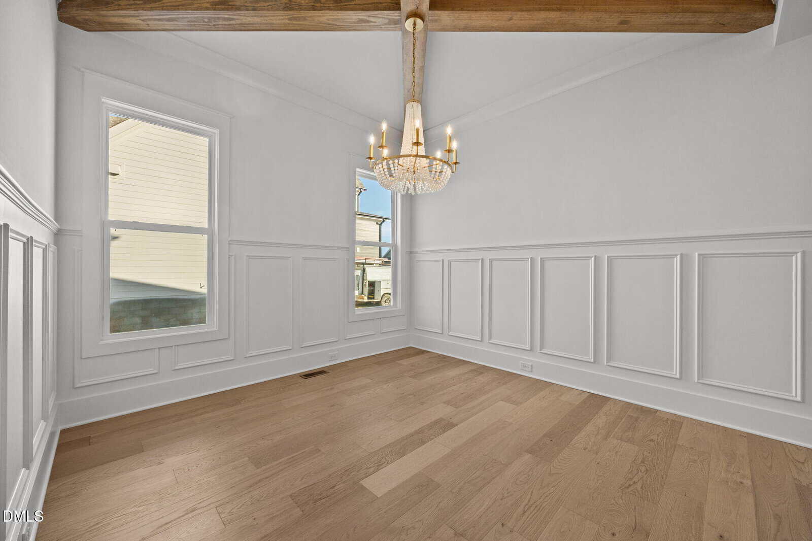 A bright, empty room with light wood floors, white wainscoting on the walls, a window, and a chandelier hanging from a ceiling with exposed wooden beams.