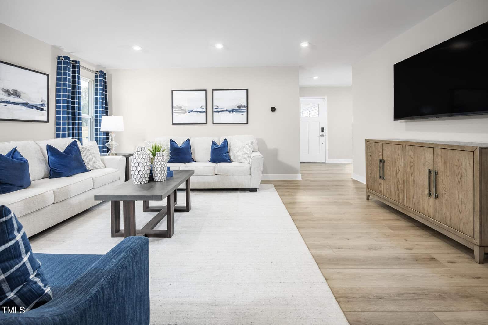 Modern living room with light-colored sofas, blue accent pillows, a blue armchair, a wooden coffee table, a large TV on the wall, artwork above the couch, and wood flooring with a light area rug.