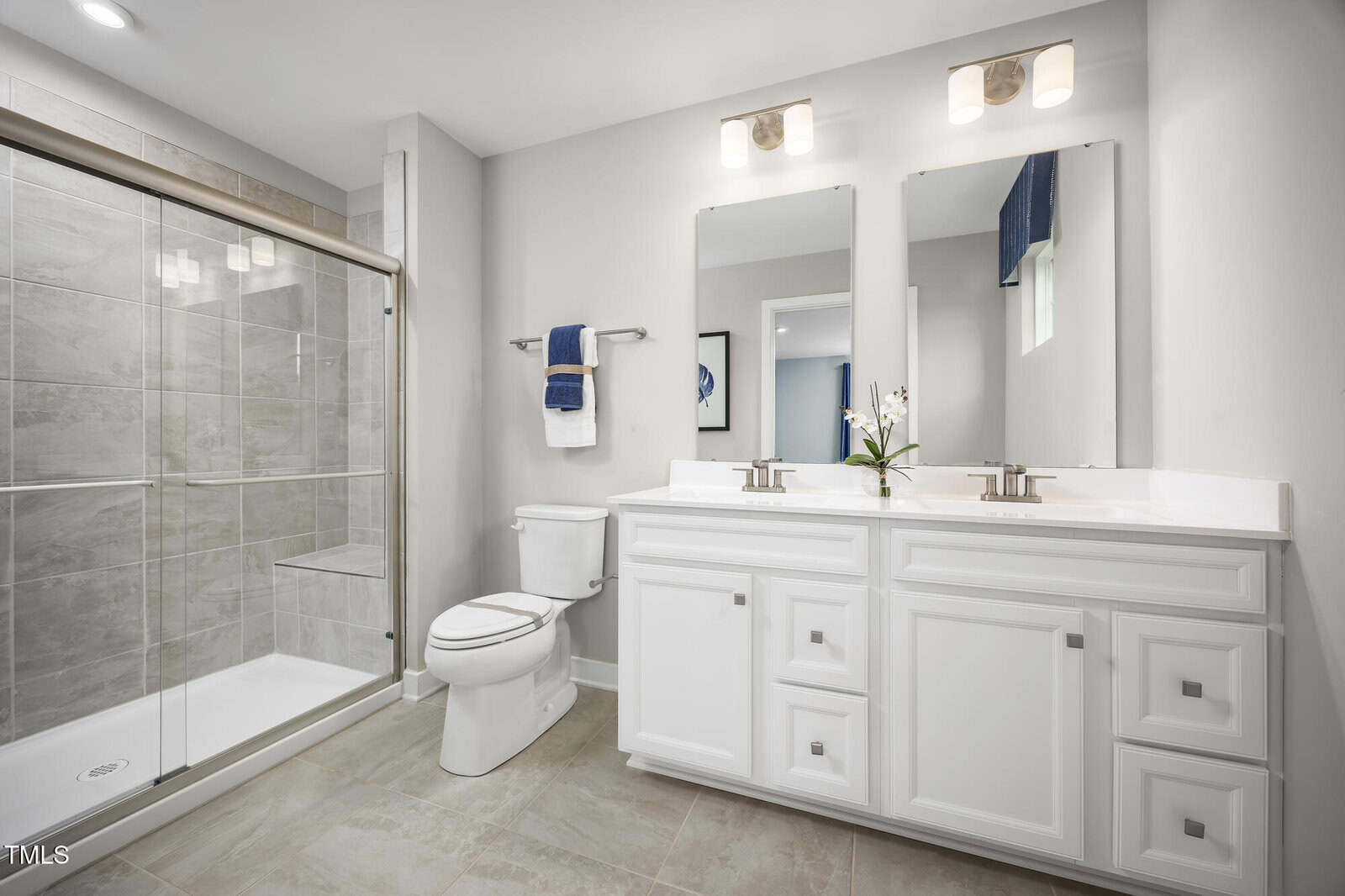Modern bathroom with a glass-enclosed shower, white vanity with dual sinks, two mirrors, a toilet, and neutral tile flooring. A towel hangs on the wall and a small potted plant sits on the counter.