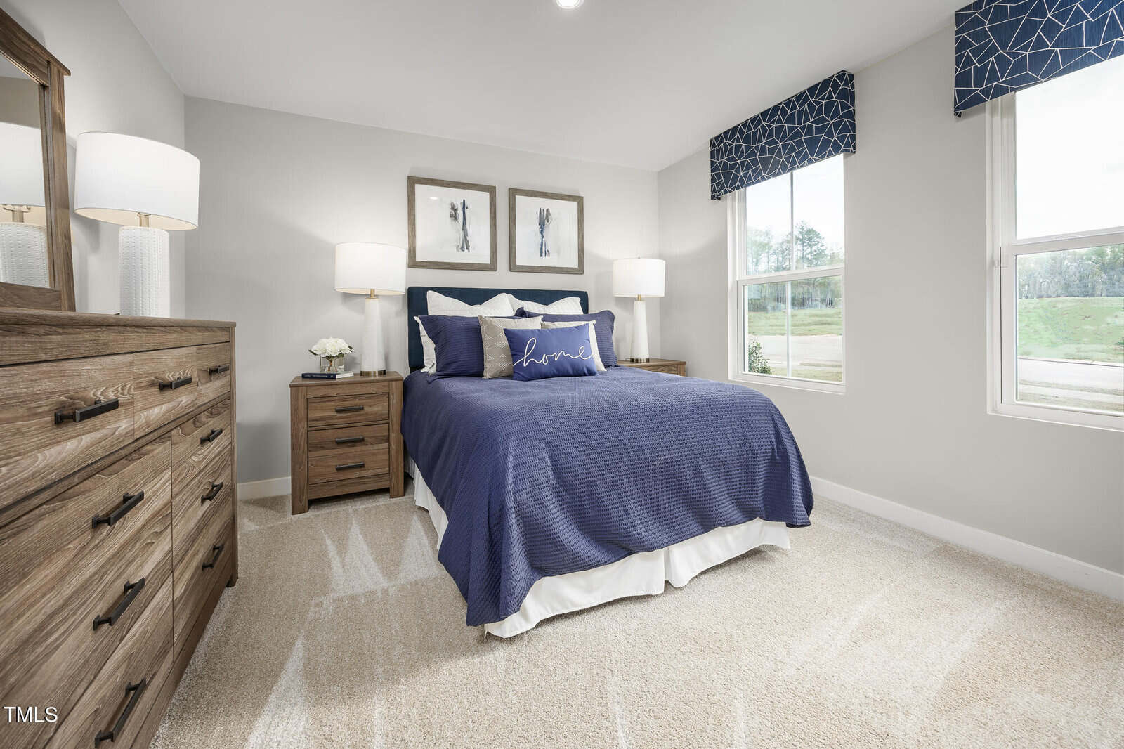 A bright bedroom with beige carpet, a wooden bed with blue bedding, flanked by matching nightstands and lamps, dressers on the left, two large windows, and three framed art pieces above the bed.