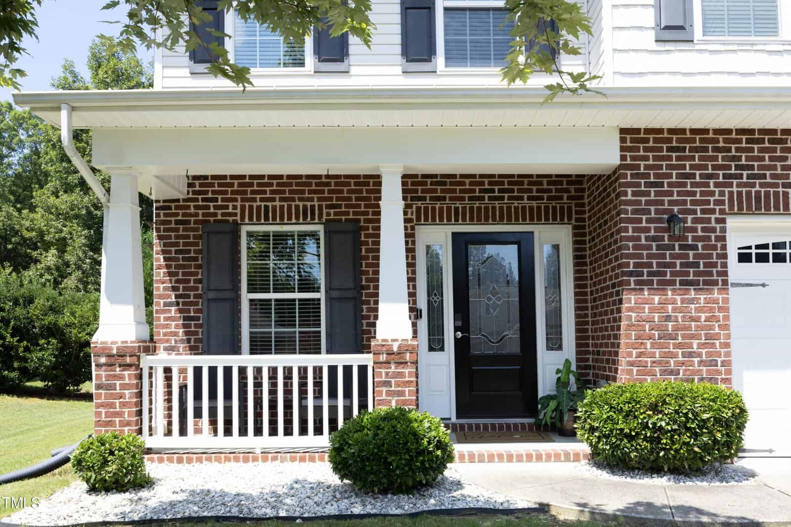 A brick house with a small front porch featuring white columns, a black front door with glass panels, two windows with dark shutters, and neatly trimmed shrubs in front.