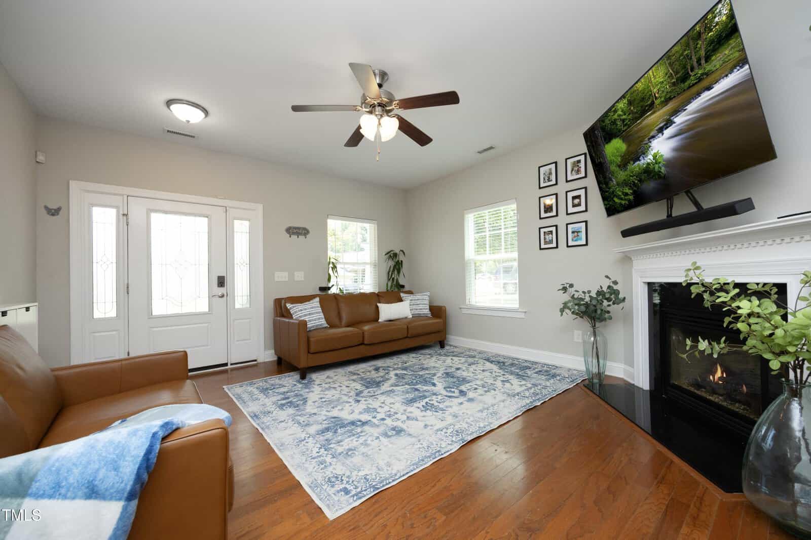A living room with brown leather sofas, a blue patterned rug, hardwood floors, a ceiling fan, a fireplace with a TV mounted above it, plants, and framed pictures on the wall near a white-framed window.