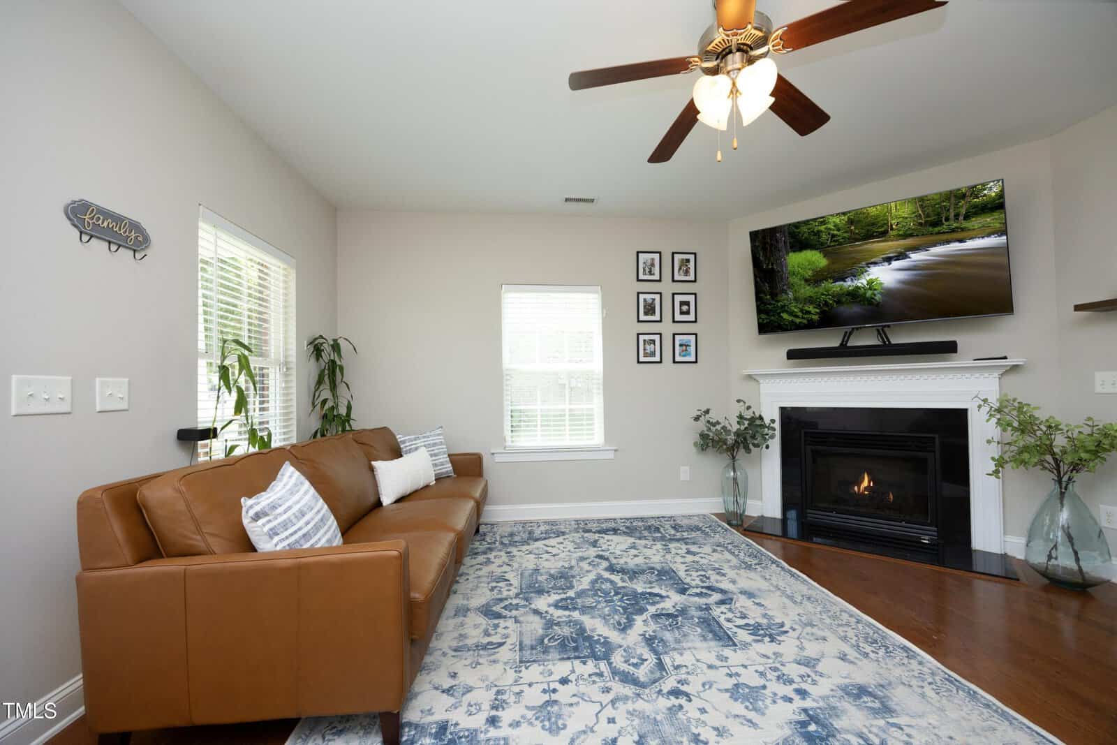 A living room with a brown leather sofa, blue and white rug, fireplace with a fire, TV mounted above showing a nature scene, wall art, and plants. A ceiling fan with lights hangs from the white ceiling.