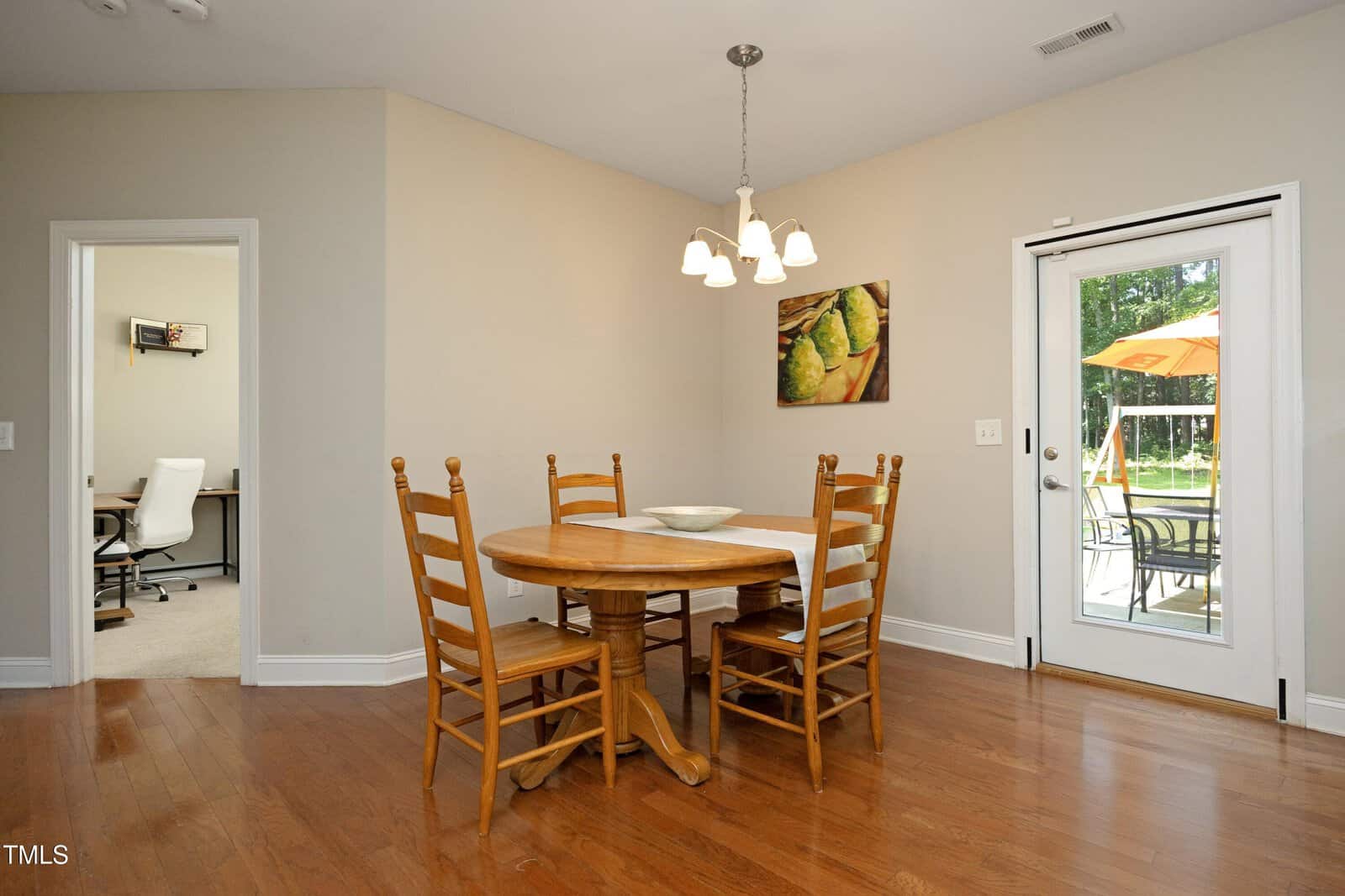 A small dining area with a round wooden table and four chairs on a hardwood floor, a chandelier overhead, a painting on the wall, a door leading outside, and an adjoining room with a desk and office chair visible.