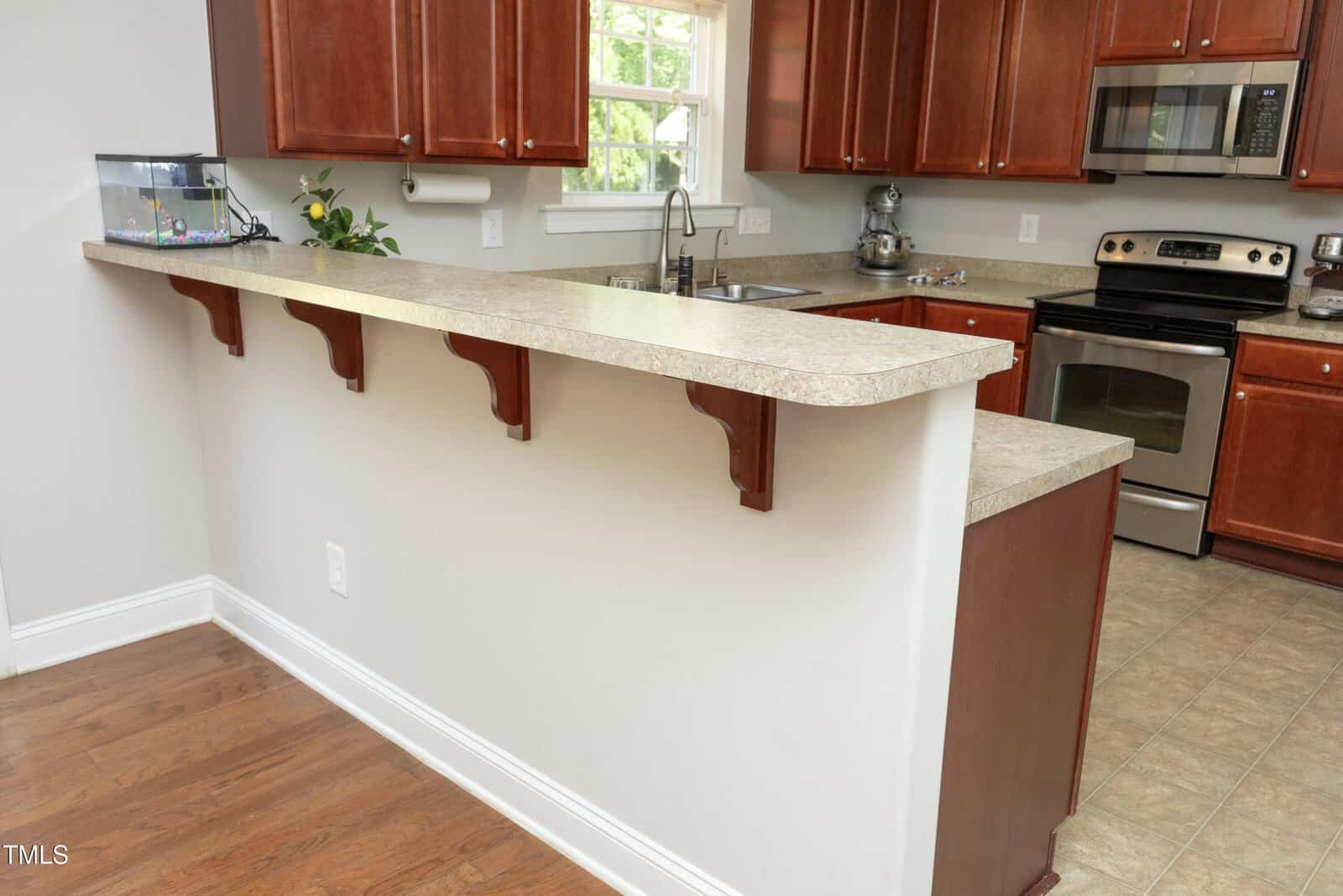 A kitchen with brown cabinets, stainless steel appliances, a double sink, tan countertops, and a raised breakfast bar. A small fish tank sits on the counter and a window is above the sink.
