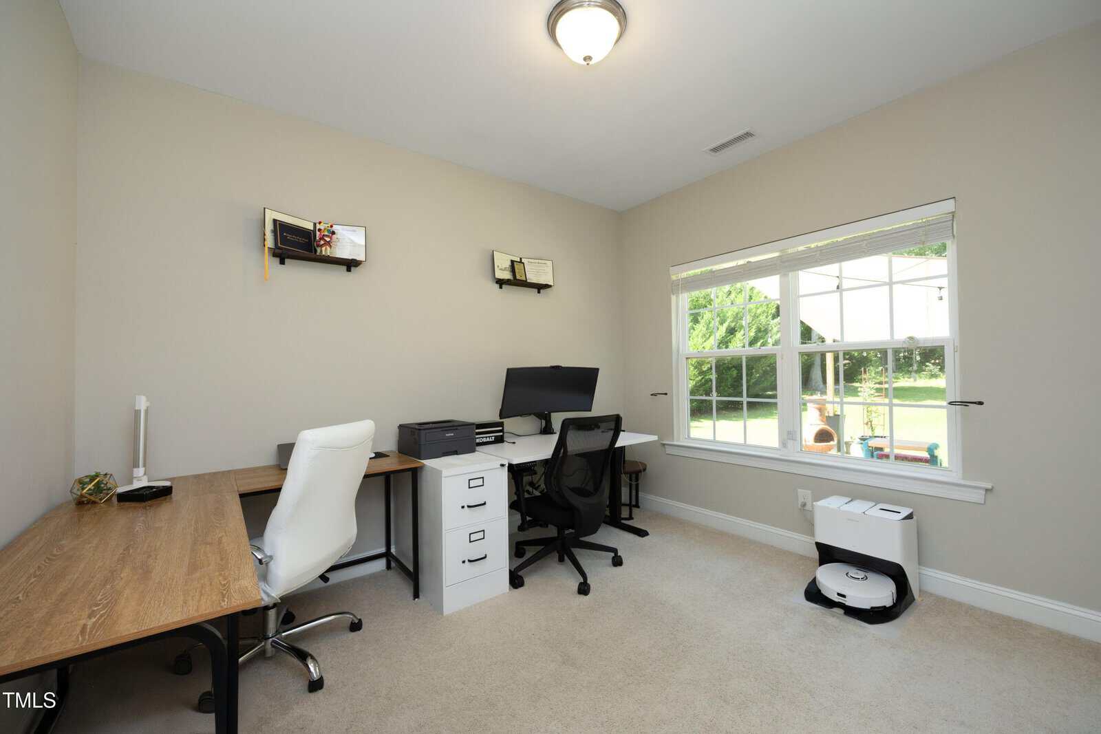A home office with two desks, office chairs, a computer monitor, shelves with items, a large window showing a sunny yard, and a robotic vacuum on the floor. The room has beige walls and carpet.