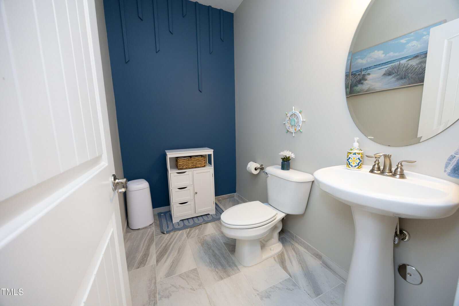 A bathroom with blue and white walls, a round mirror, pedestal sink, toilet, small cabinet, trash bin, and beach-themed decor. Blue accent wall and tile floor are visible.