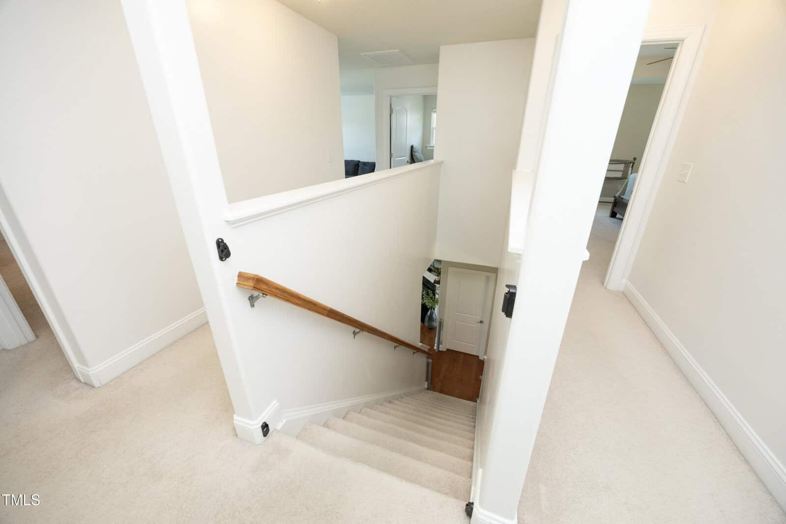 Carpeted staircase with a wooden handrail leads down from a hallway with white walls and doors on both sides; a small area with wood flooring is visible at the bottom.