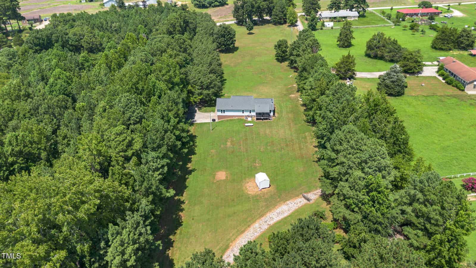 Aerial view of a house surrounded by trees, with a large grassy yard, a small shed near the center, and a gravel driveway leading to the house. Several neighboring houses are visible in the distance.
