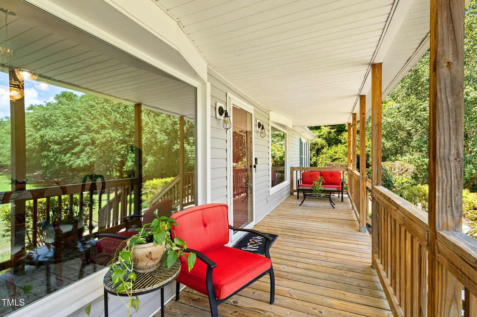 A wooden front porch with a white ceiling, two red cushioned chairs, a small round table with a potted plant, and wooden railings. Large windows reflect trees and greenery from the yard.