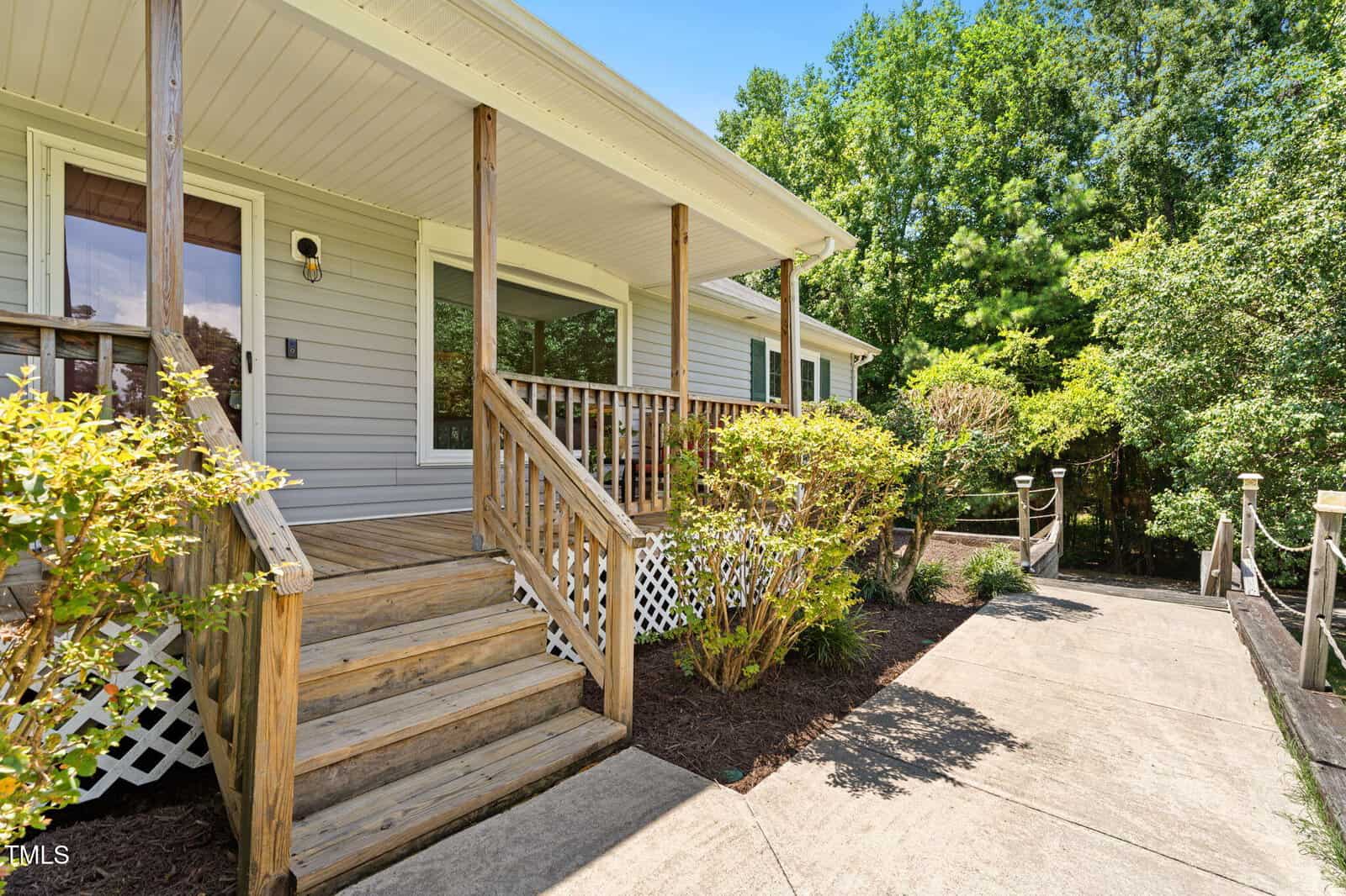 A wooden porch with steps and railing leads to the entrance of a light gray house. Shrubs and greenery line the walkway and the house is surrounded by trees under a clear sky.