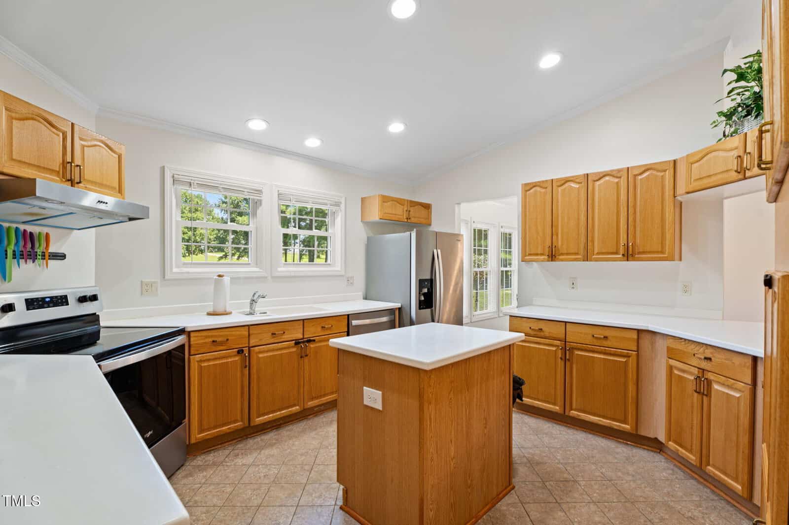 A spacious kitchen with wooden cabinets, white countertops, a stainless steel refrigerator, an electric stove, an island in the center, and multiple windows letting in natural light.