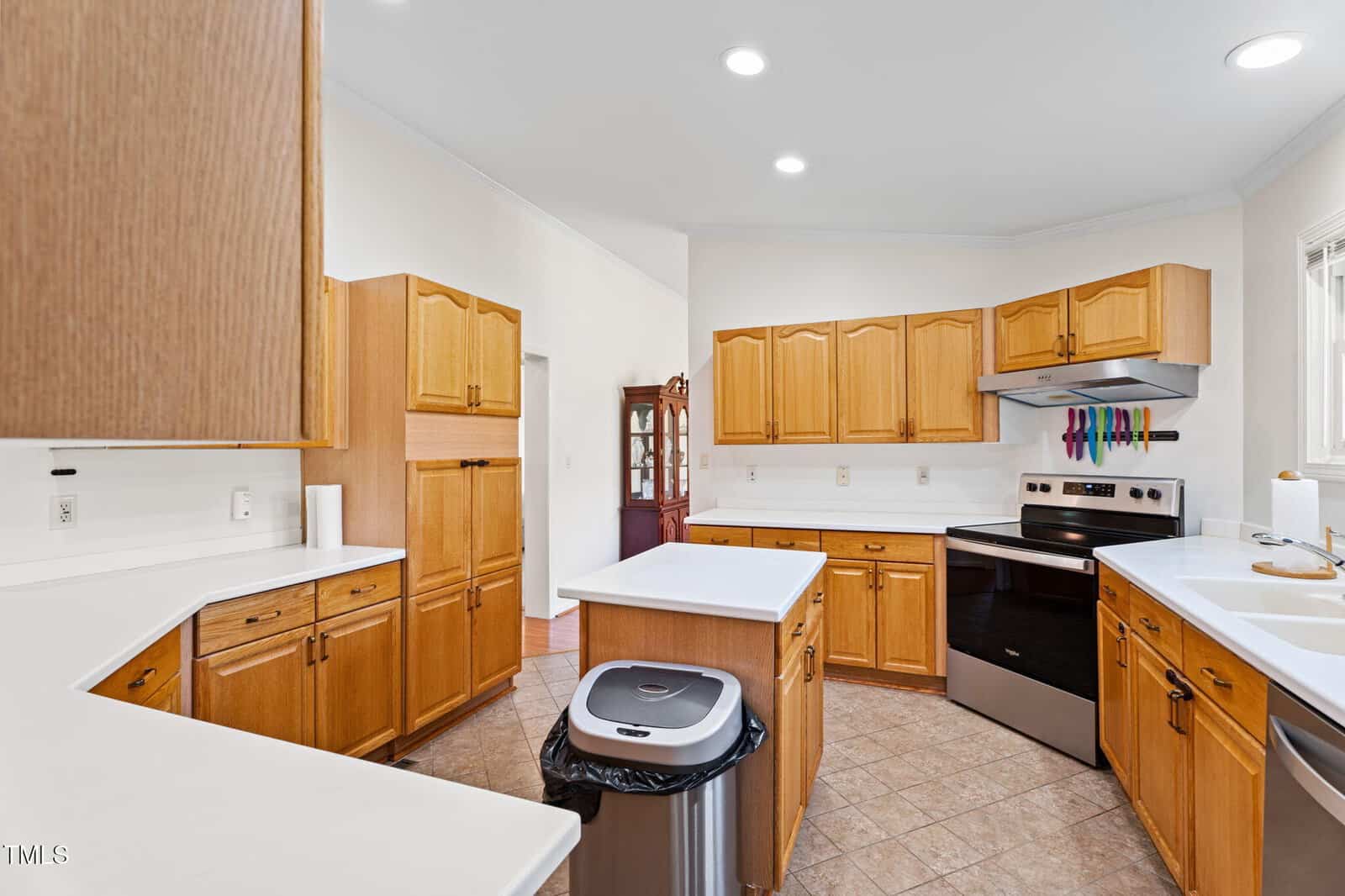 Spacious kitchen with light wood cabinets, white countertops, stainless steel appliances, a trash can in the center, tile flooring, and recessed ceiling lights.