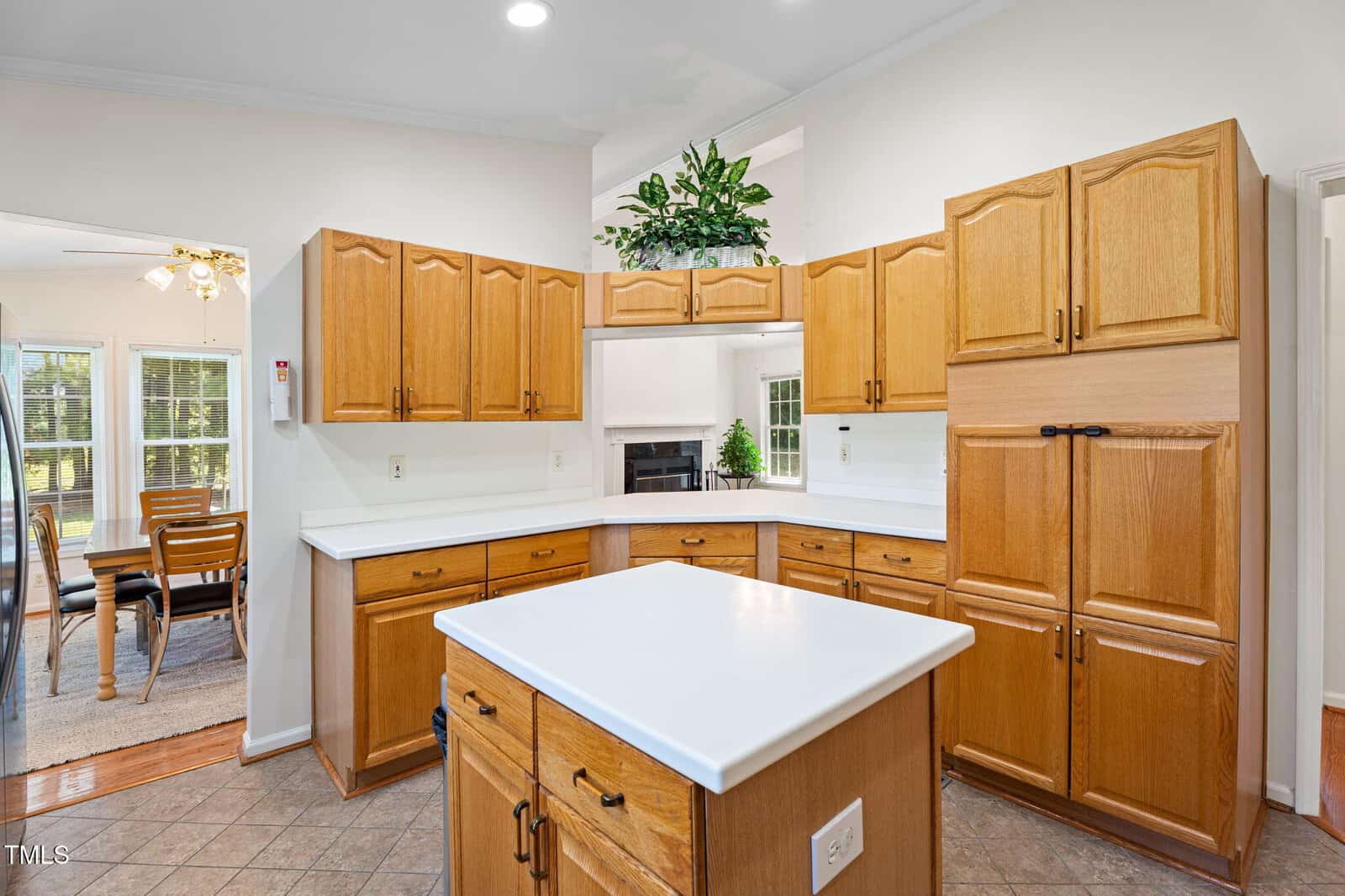 Spacious kitchen with light wood cabinets, white countertops, and a center island. Dining area with wooden table and chairs is visible through an open doorway. Large windows let in natural light.