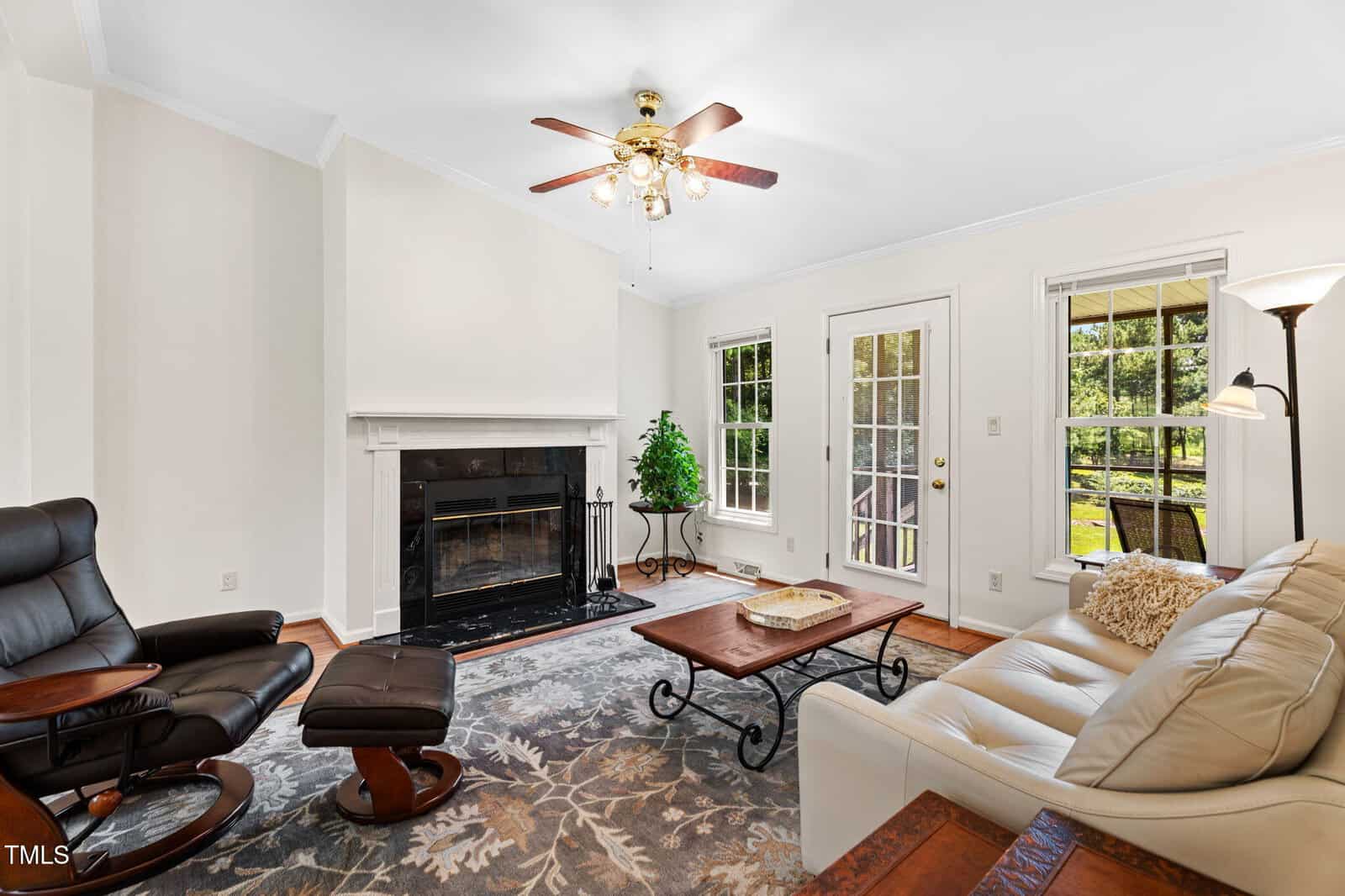 A living room with a cream-colored sofa, a dark recliner with footrest, a coffee table, a fireplace, and a floor lamp. Large windows and a glass door offer a view of a green yard outside.