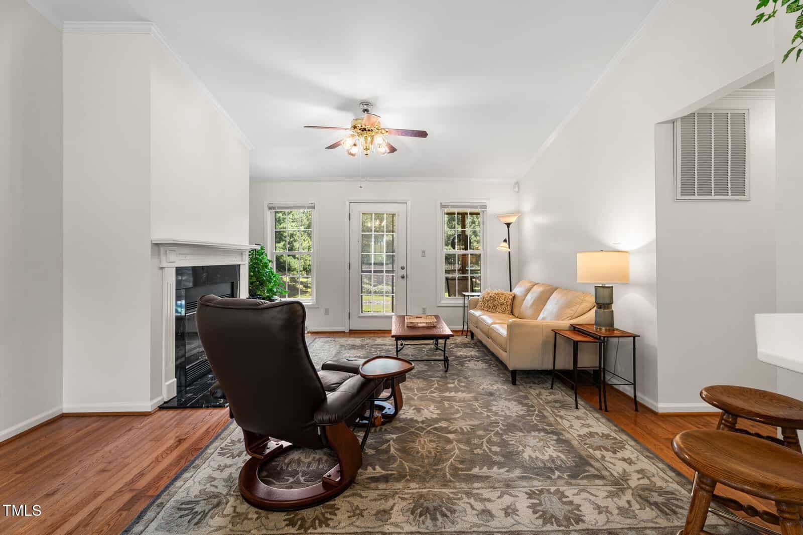 Living room with hardwood floors, a patterned rug, a brown recliner, a beige sofa, a coffee table, and a floor lamp. French doors and windows reveal greenery outside. Ceiling fan and fireplace are also visible.