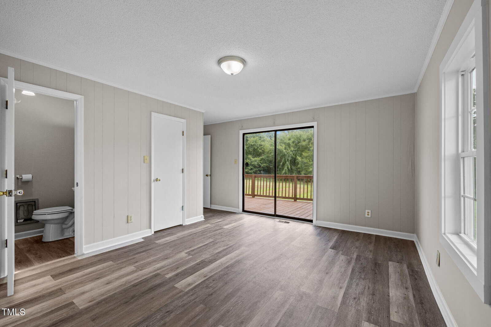 Bright, empty room with wood flooring, light-colored walls, a ceiling light, sliding glass door to a deck, windows on the right, and an open door to a bathroom on the left.