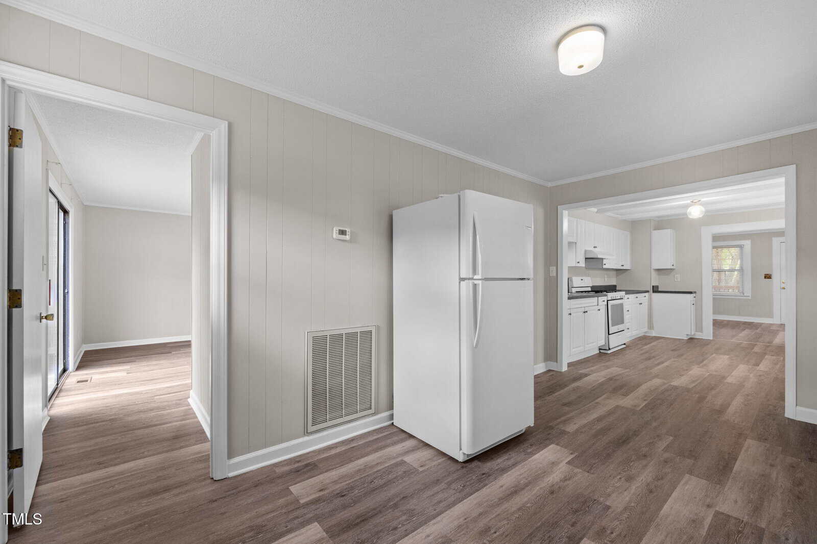 A kitchen with white cabinets and appliances, wood-look flooring, and beige paneled walls. An adjacent dining area and another room are visible through open doorways. Ceiling lights are turned on.