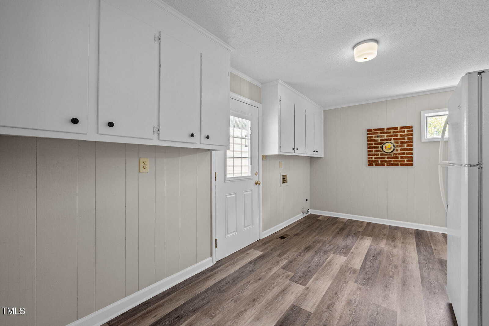 A kitchen with white upper cabinets, light wood-paneled walls, dark wood flooring, a white door with a window, a small window, a refrigerator, and a brick accent wall with a round clock.