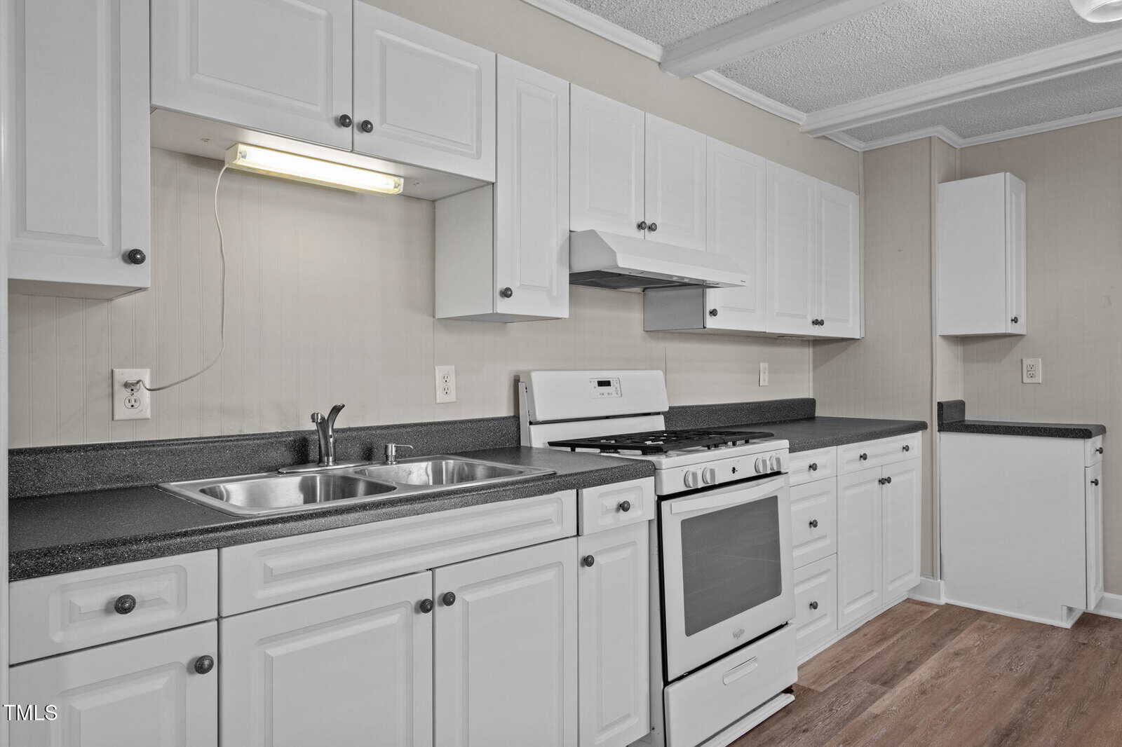 A kitchen with white cabinets, black countertops, a double sink, gas stove with oven, range hood, and wood-style flooring. There are multiple drawers and cabinets along the wall, with under-cabinet lighting above the sink.