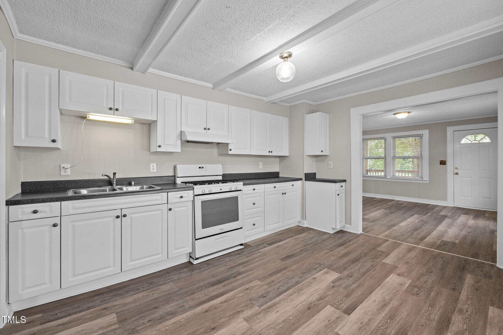 A kitchen with white cabinets, a white stove, and dark countertops. The floor is wood laminate, and the walls are light-colored. An open doorway leads to a living area with windows and a white front door.