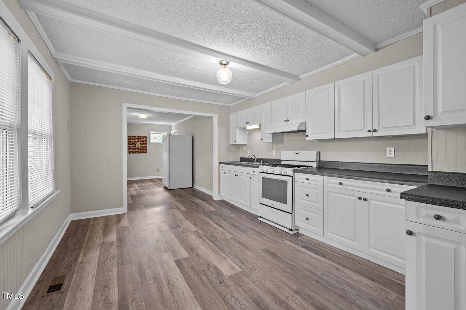 Bright kitchen with white cabinets, white appliances, and gray countertops. Wood-look vinyl flooring runs throughout. Large windows on the left let in natural light. The kitchen opens to another room at the back.