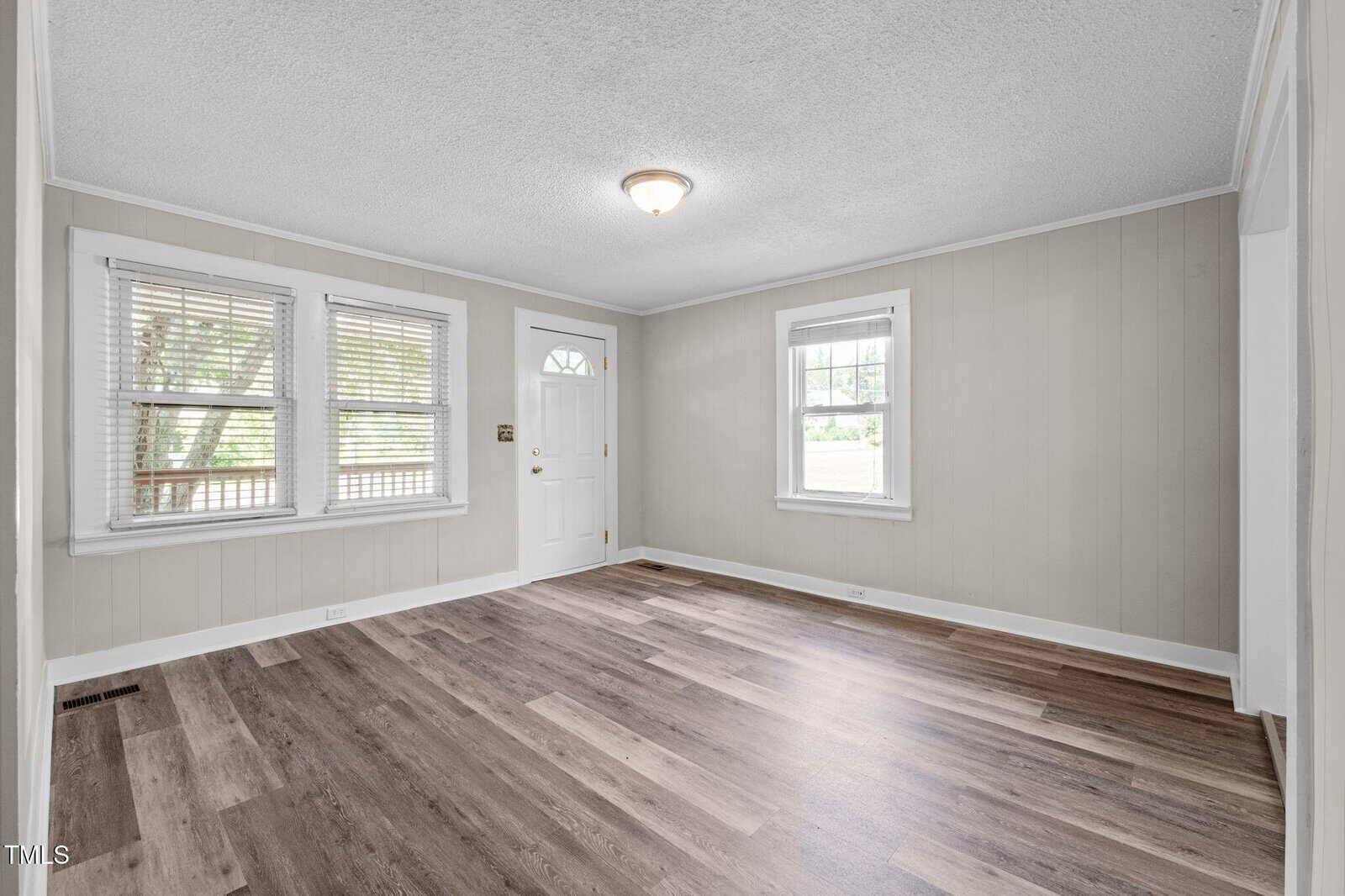 An empty room with light-colored walls, a white front door, three windows letting in natural light, a ceiling light fixture, and wood-style flooring in various shades of brown.