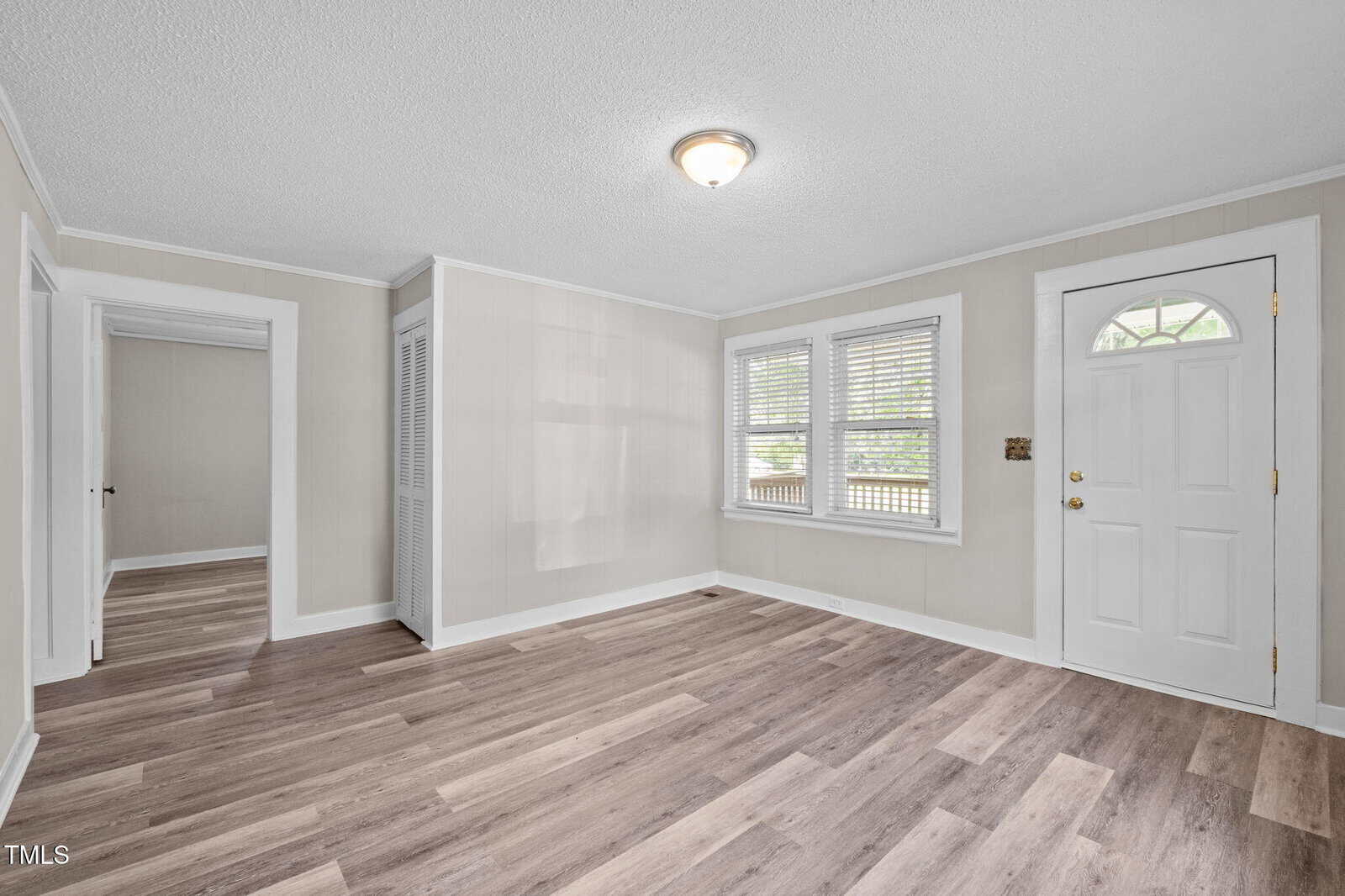 A bright, empty room with light beige walls, wood-patterned vinyl flooring, two windows with blinds, a white front door with a window, and visible doorways to other rooms. Ceiling light fixture is on.