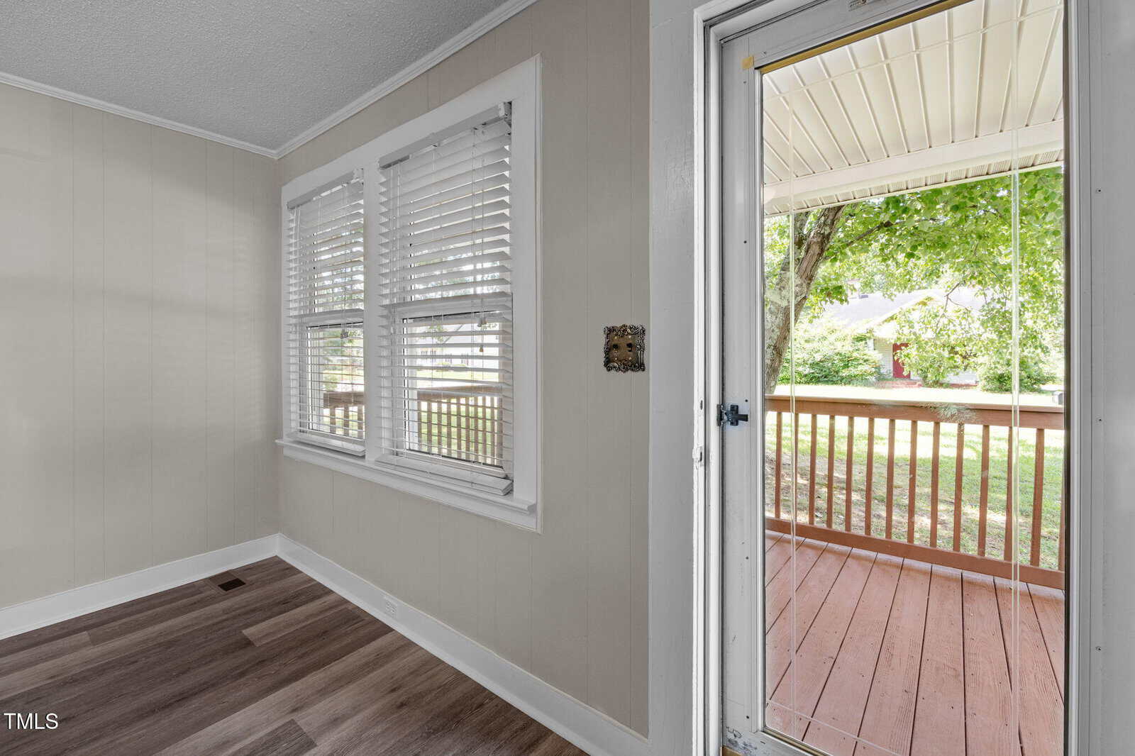 A small room with light-colored walls and wood-style flooring features two windows with white blinds and an open door leading to a covered wooden porch with a railing outside. Trees are visible in the background.