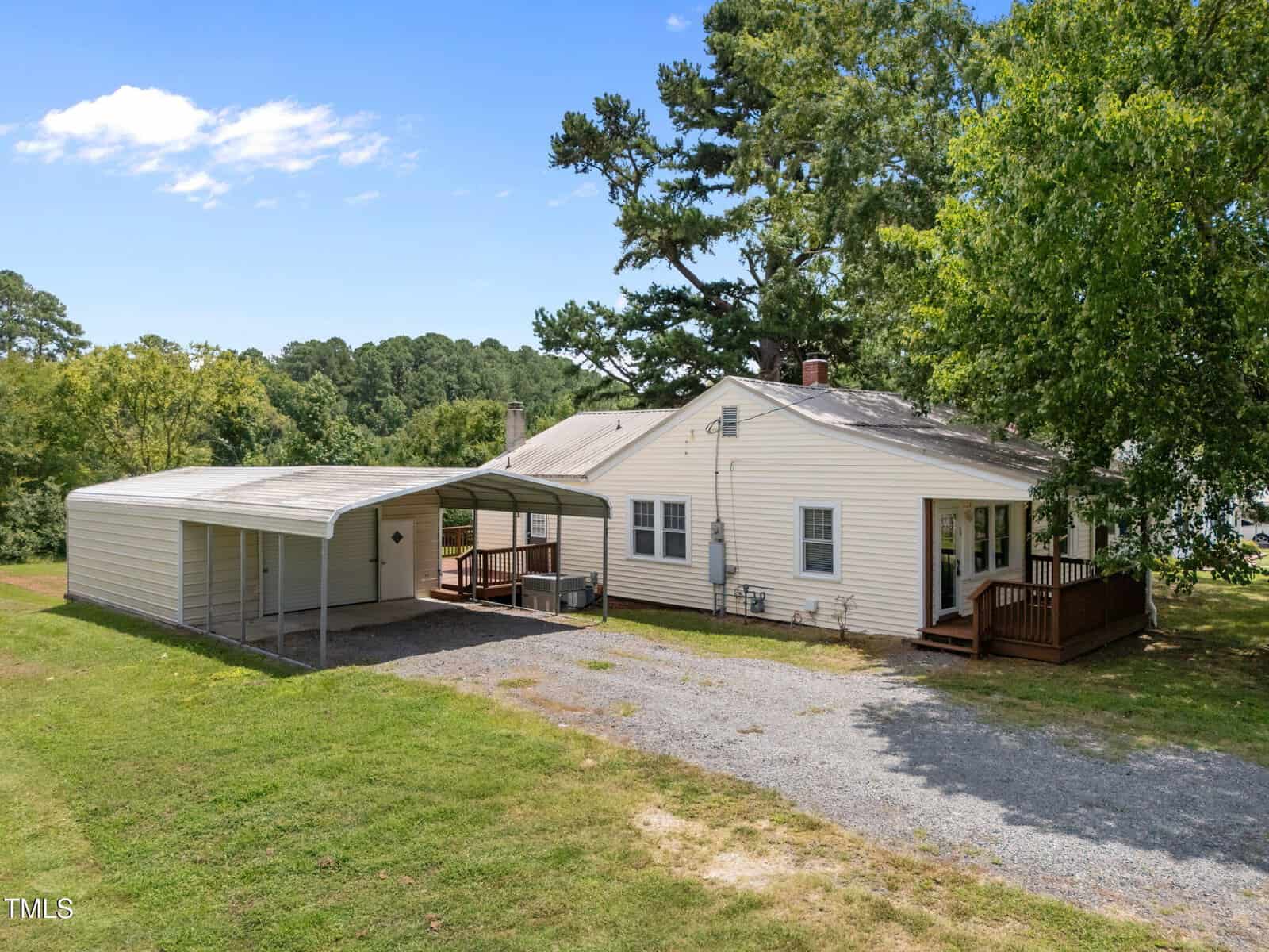 A single-story beige house with a brown wooden deck sits beside a large metal carport with two parking spaces. The house is surrounded by grass, trees, and a gravel driveway under a blue sky with some clouds.