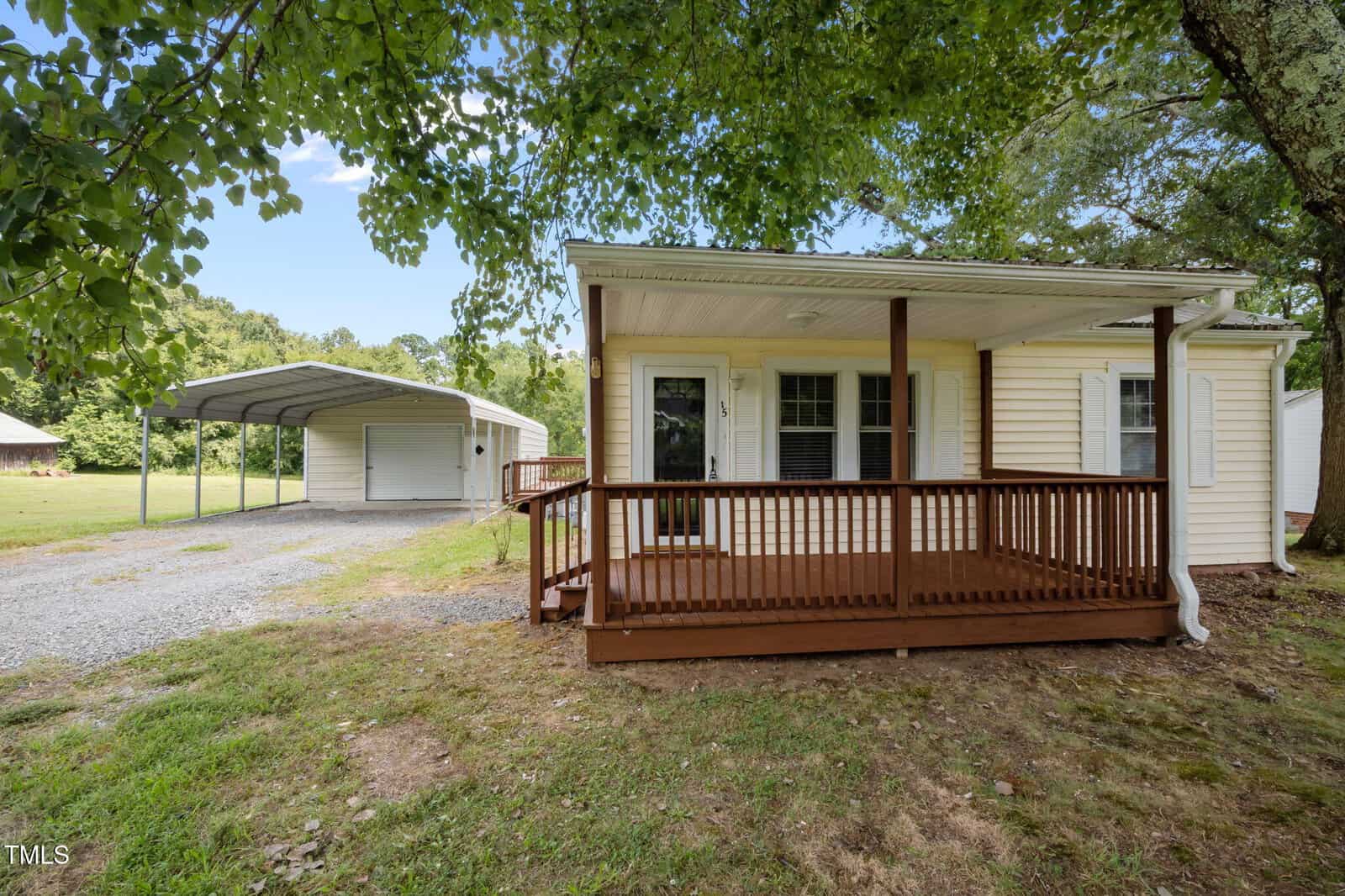 Charming single-story house at 115 Brogden Road in Stem with a covered front porch, beige siding, and white trim. A gravel driveway leads to a detached carport and storage building, all surrounded by lush trees and grass.